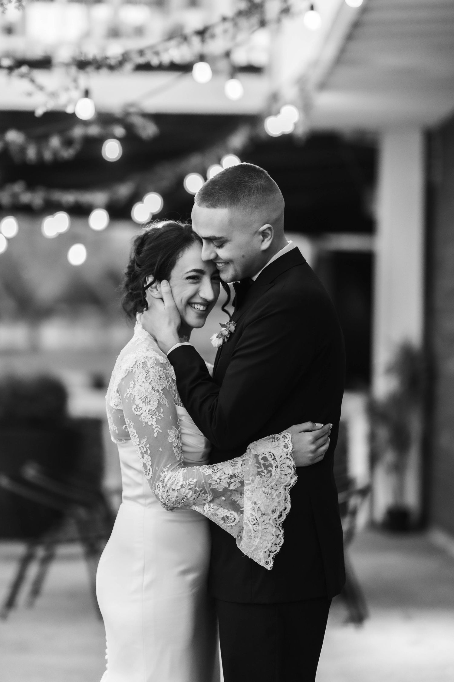 Black and white photo of a smiling bride in lace gown sharing first dance with groom in tuxedo under string lights.