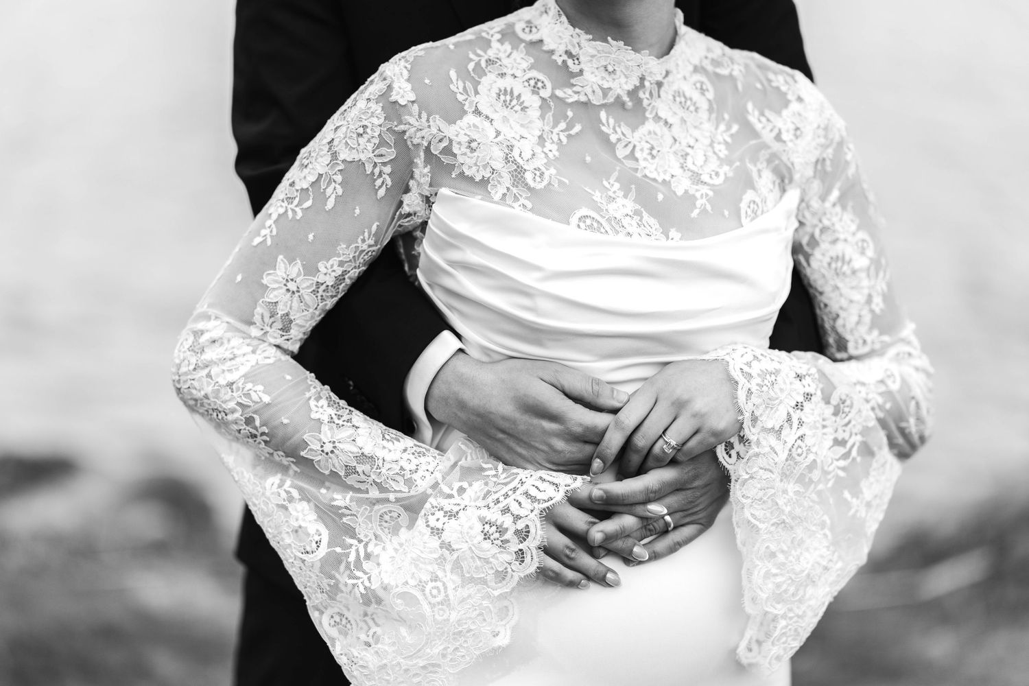 Black and white photo of a bride in lace wedding gown embraced by groom in suit.