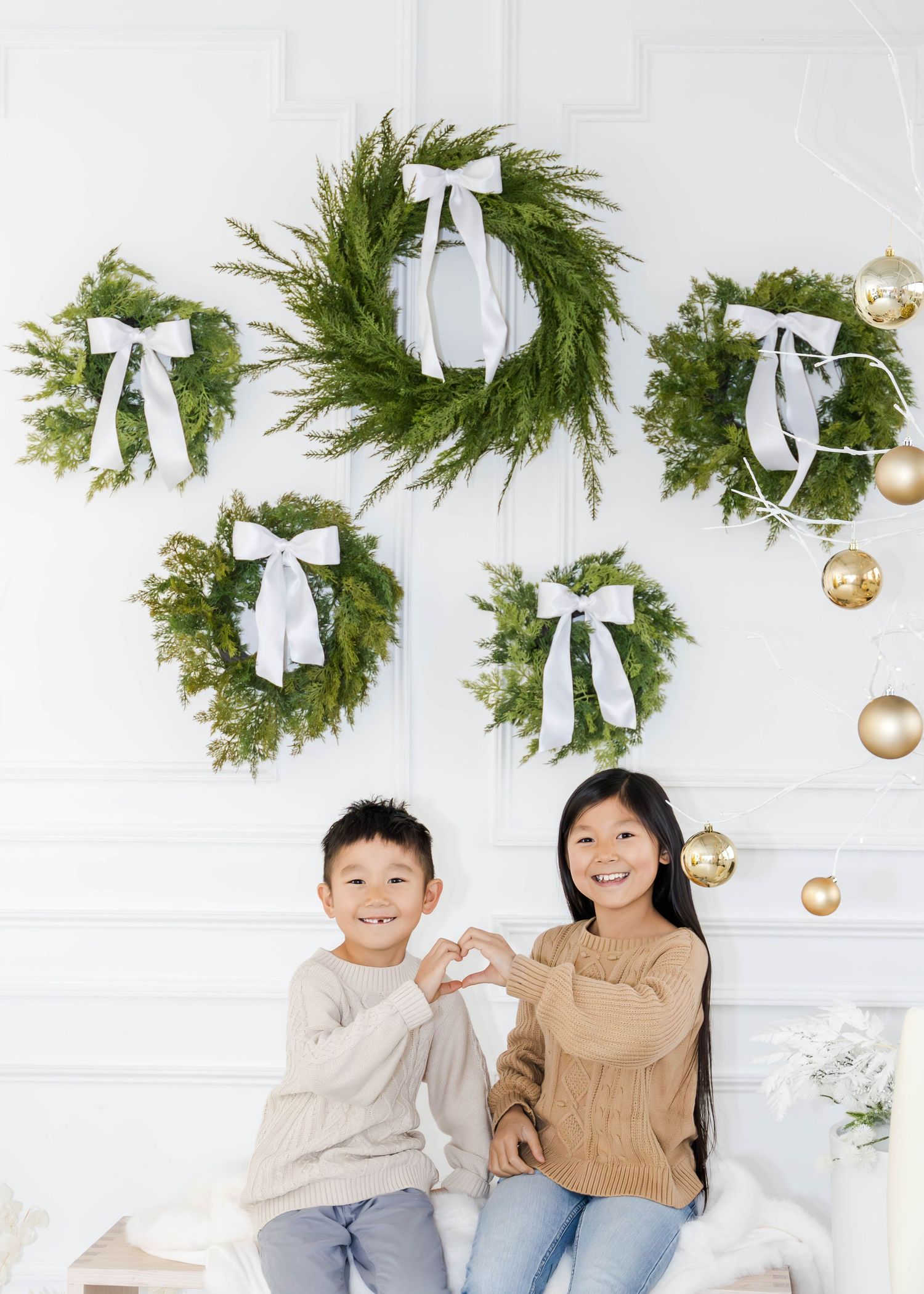 Family wearing neutral colors poses in front of Christmas wreaths with white bows and gold ornaments.