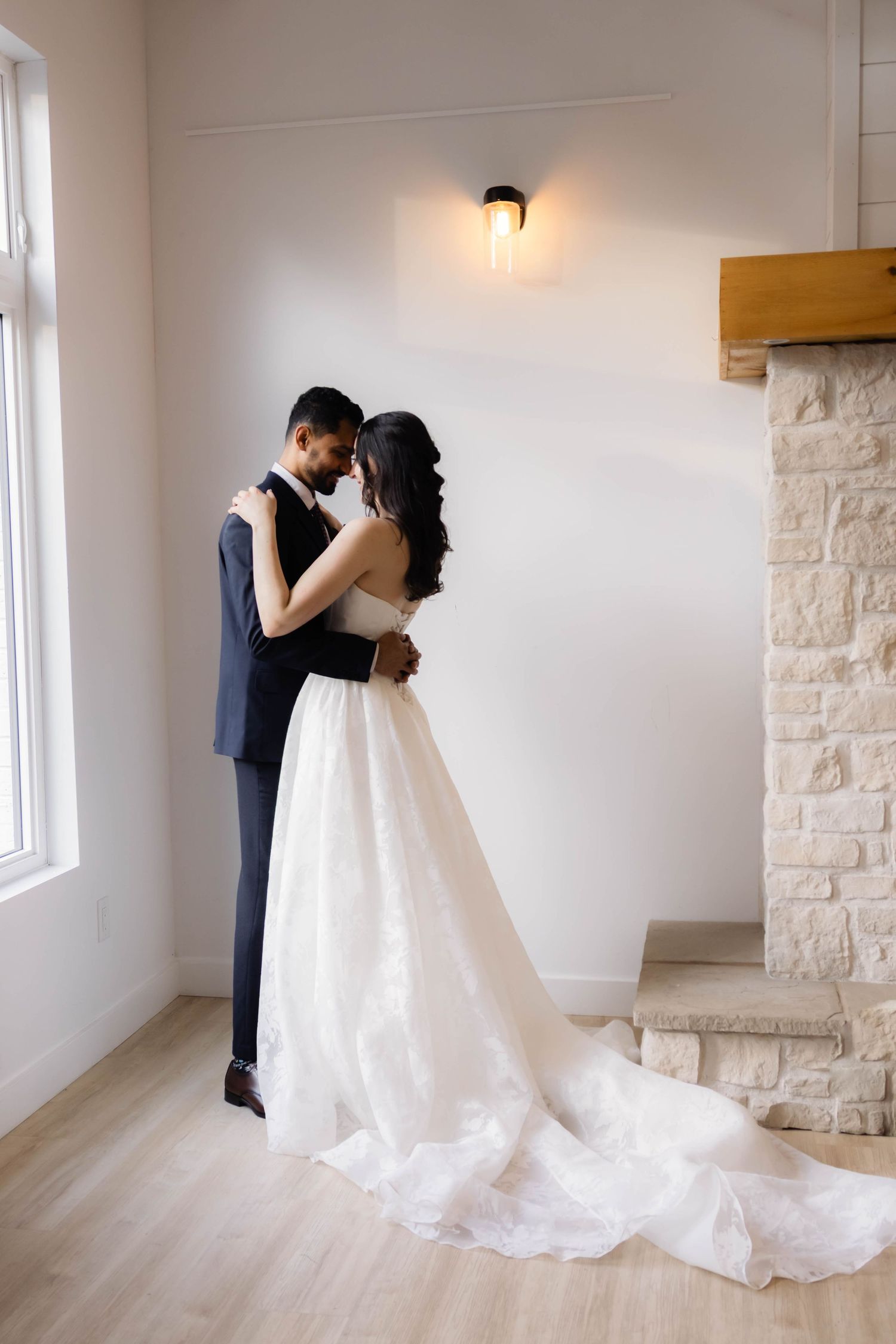 Wedding couple embracing in minimalist white room with stone accent wall.
