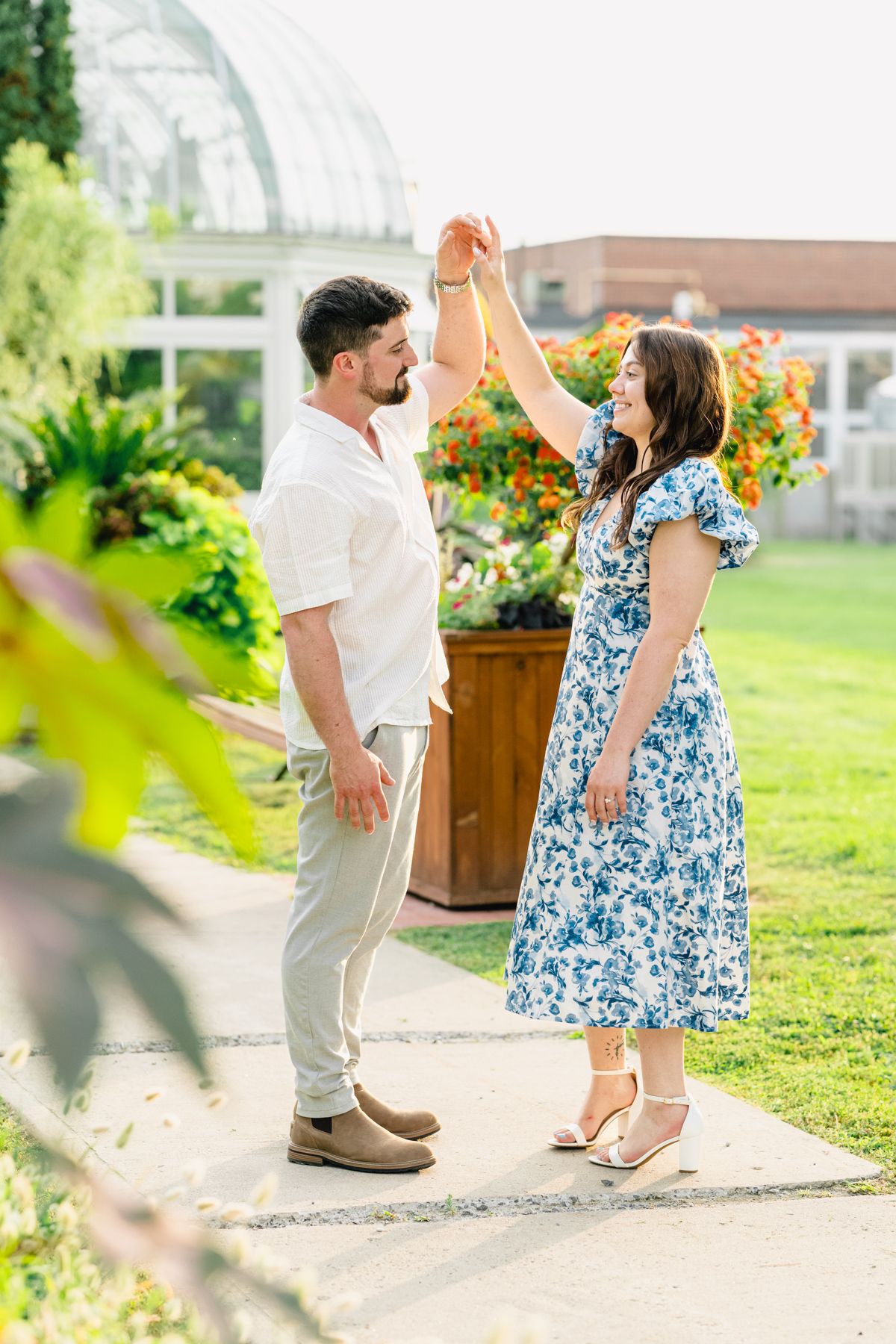 A couple dances outdoors in a garden, woman in blue floral dress, man in white shirt, surrounded by colorful flowers.
