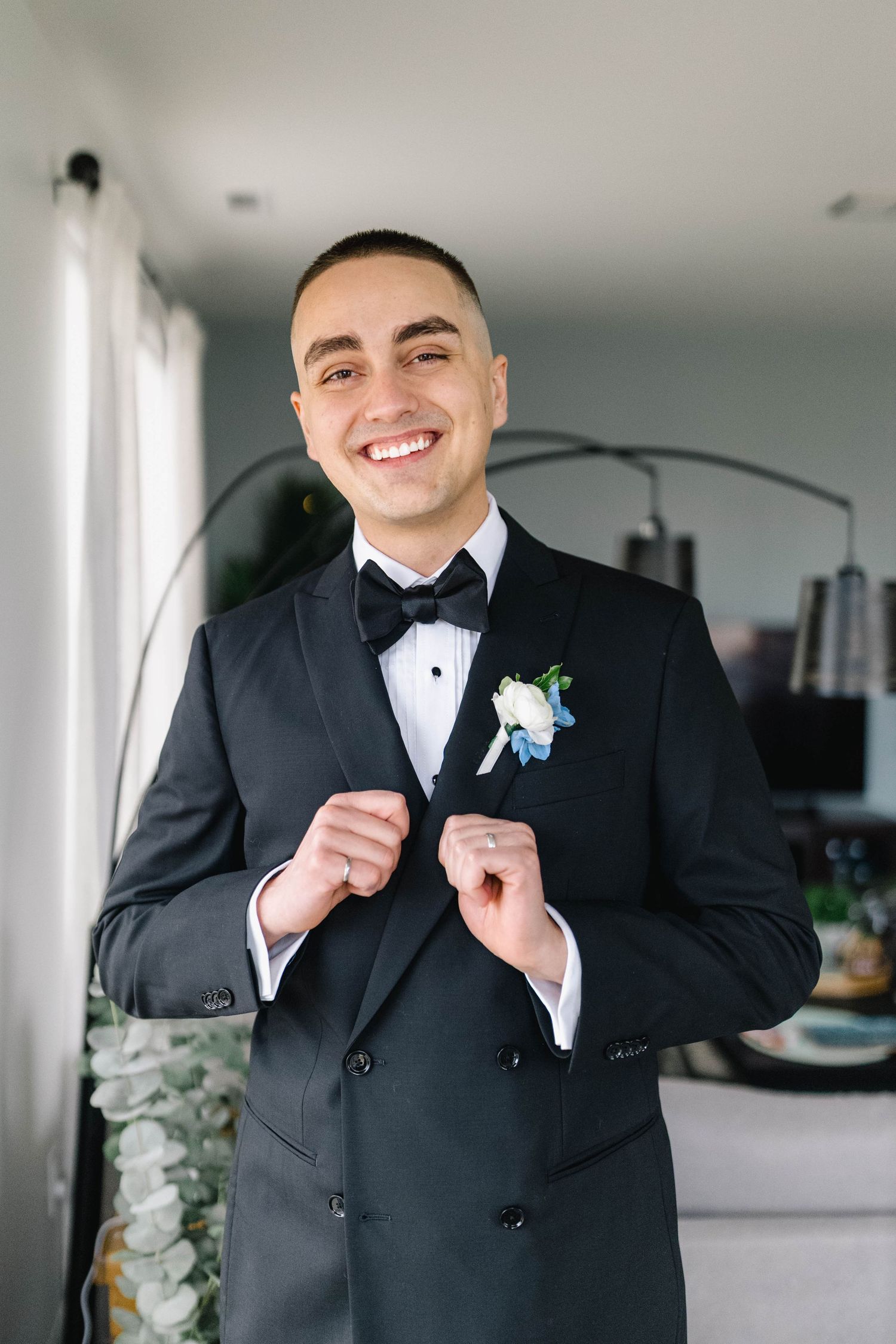 Smiling groom in black tuxedo with bow tie and white boutonniere adjusting jacket buttons indoors.