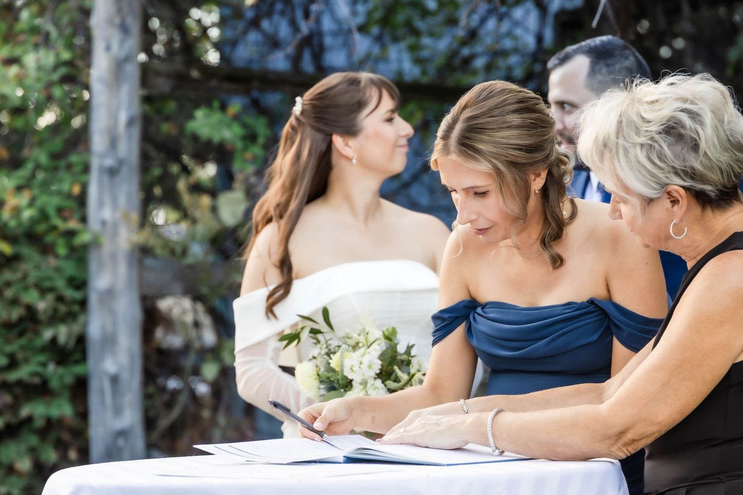 Wedding party gathers around to witness the signing of marriage documents outdoors.