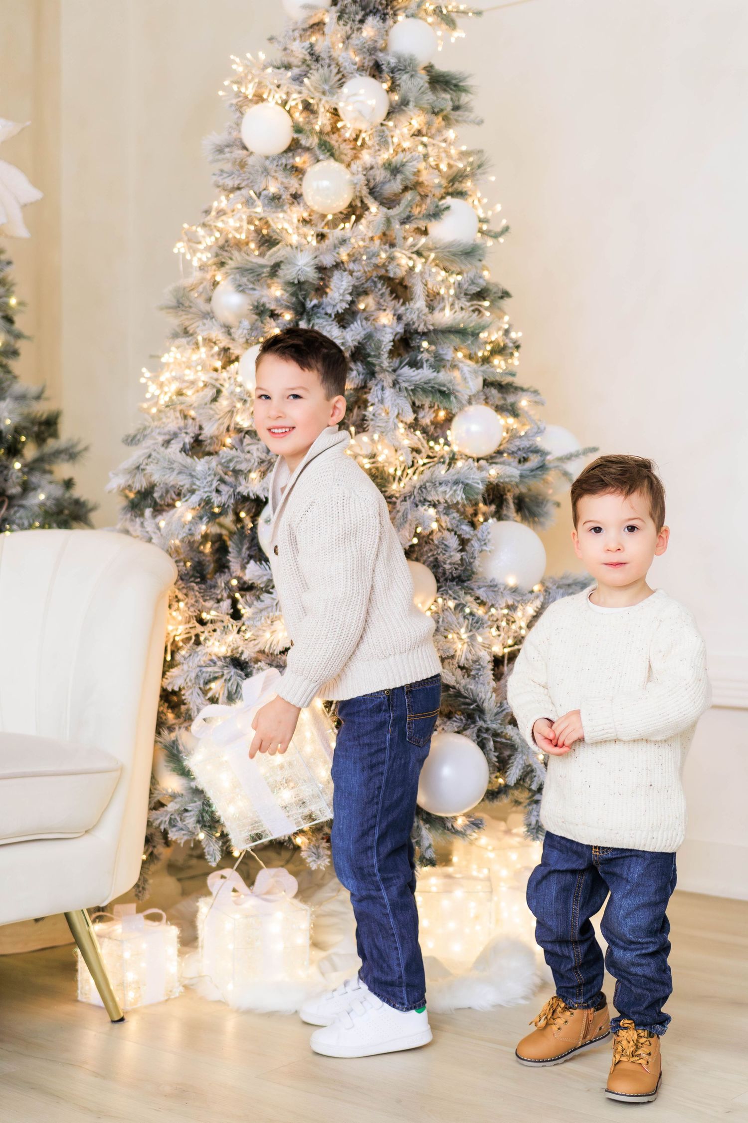 Two children in white sweaters playing near a decorated Christmas tree.