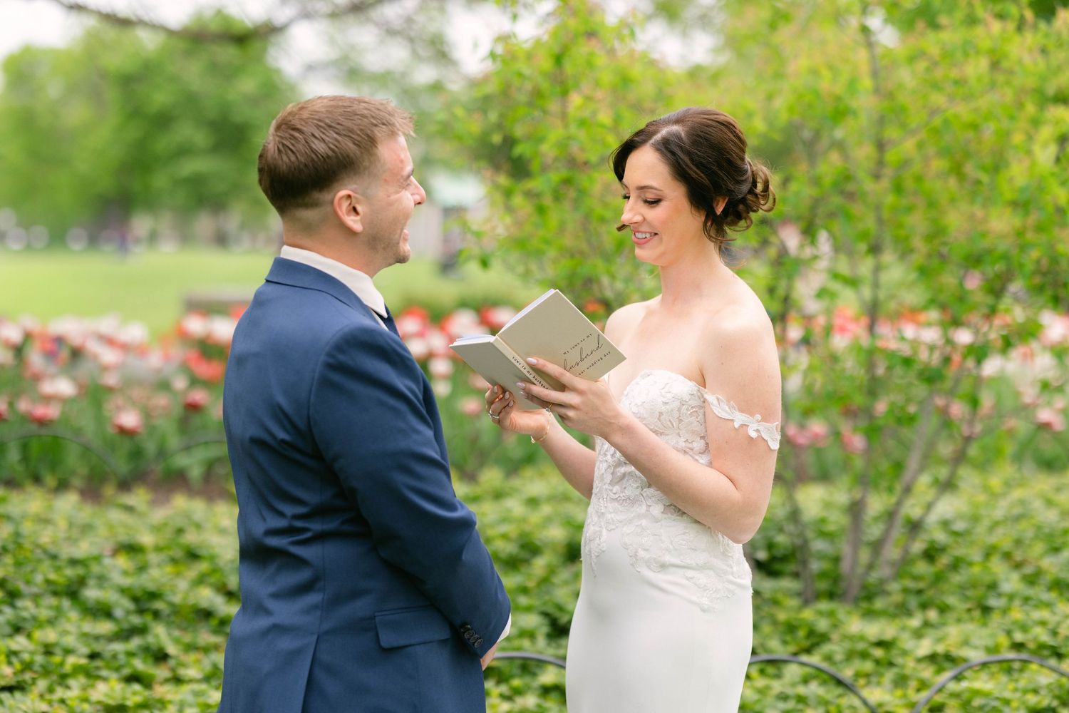 Bride in white strapless gown reads vows from book to groom in navy suit outdoors with pink flowers.