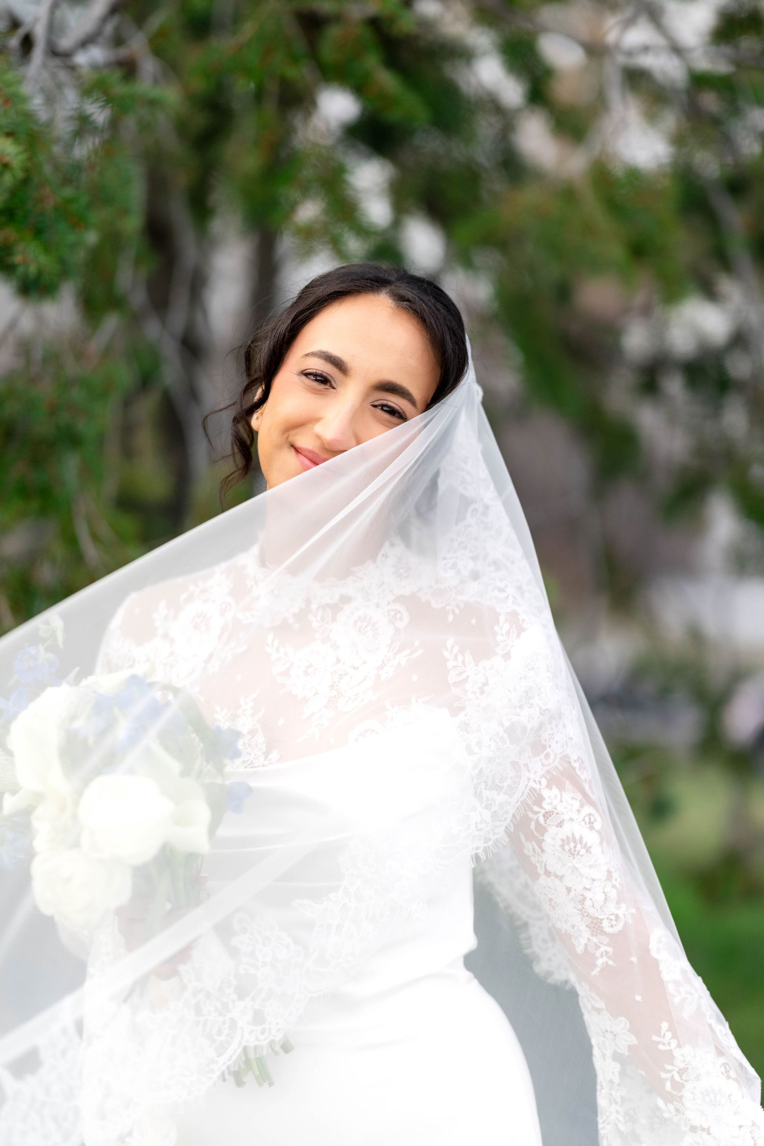 Radiant bride smiles as her lace-trimmed veil blows across her face while holding white bouquet outdoors.