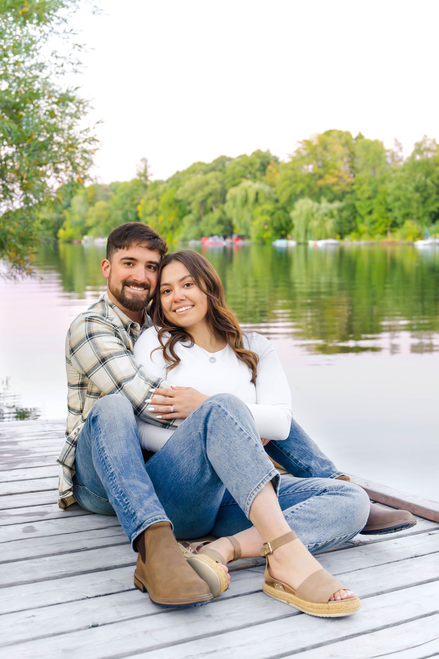 Couple sits together on wooden dock with scenic lake and lush greenery in background.