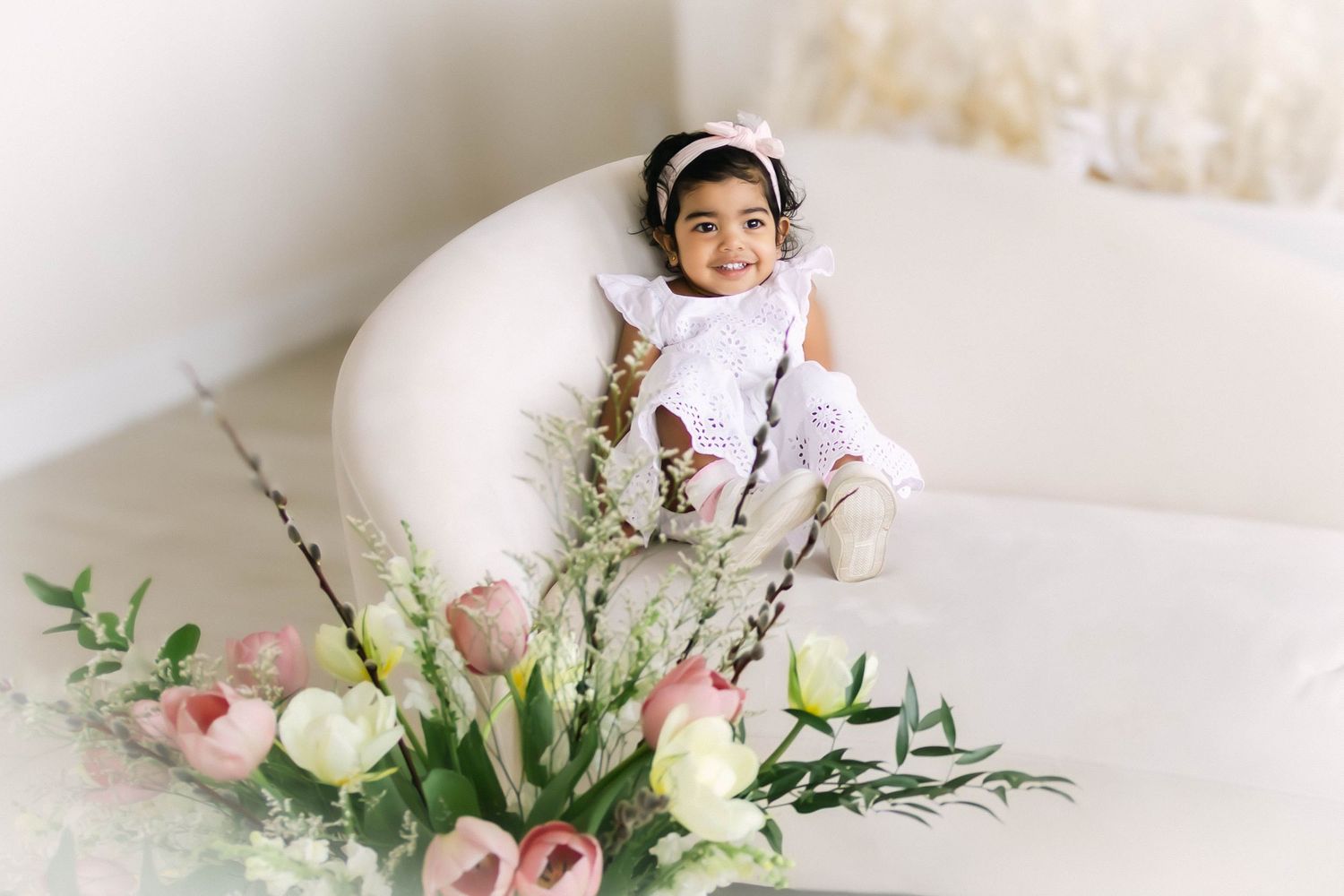 A child wearing white sits on a cream colored couch with a beautiful floral arrangement of roses and greenery in the foreground.