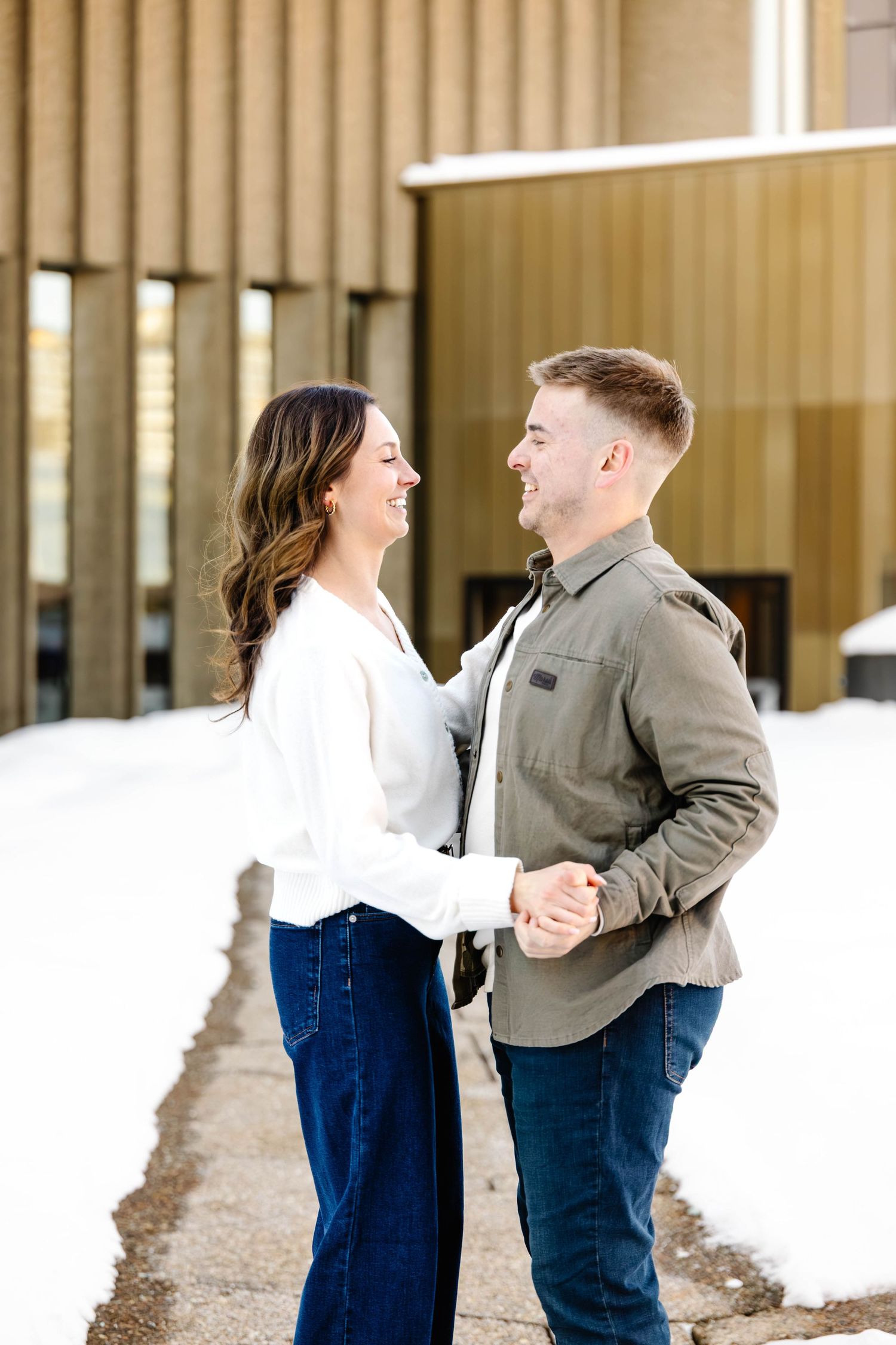 A couple in casual attire shares a romantic moment in front of a modern building with large windows.