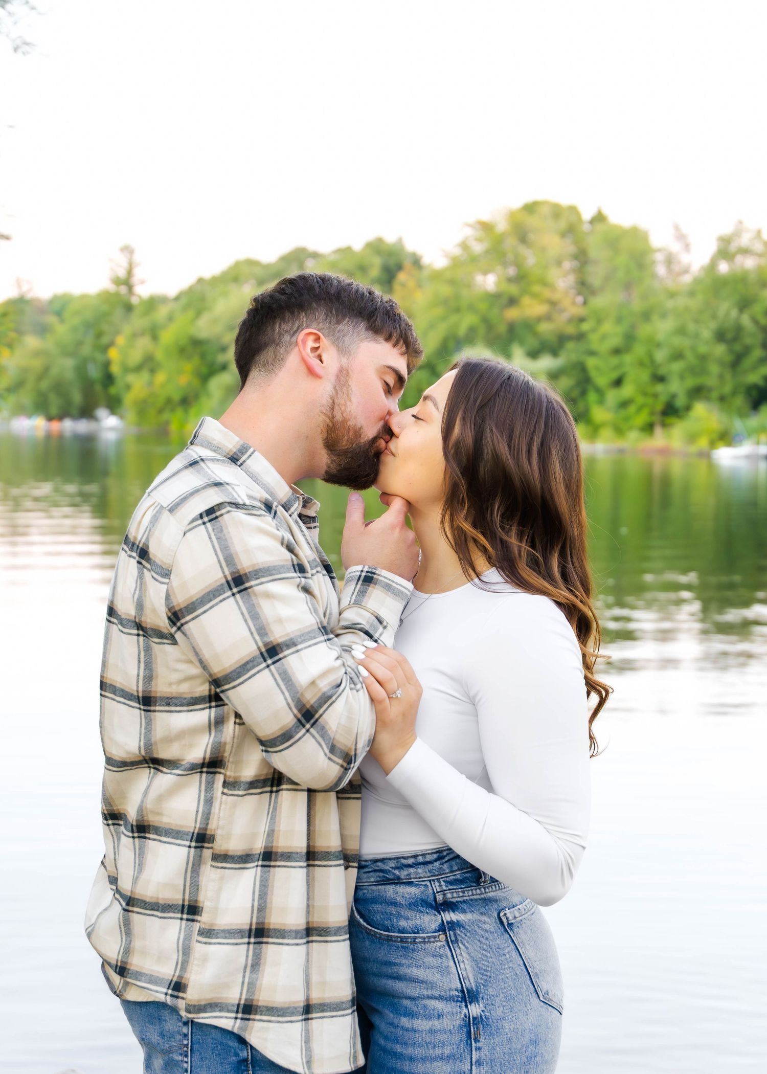 A couple in casual attire shares romantic moments by a serene lake during a sunset photoshoot.
