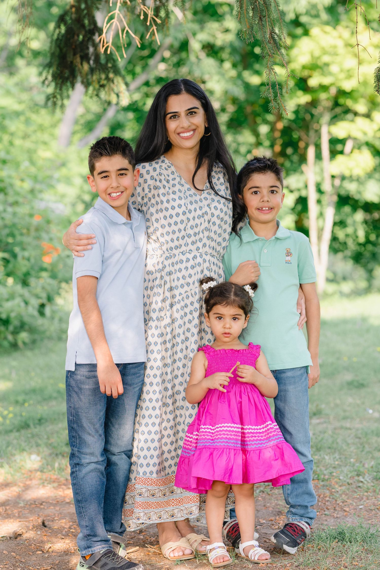 A smiling mother in a floral dress poses outdoors with her three young children, two boys and a girl in pink.
