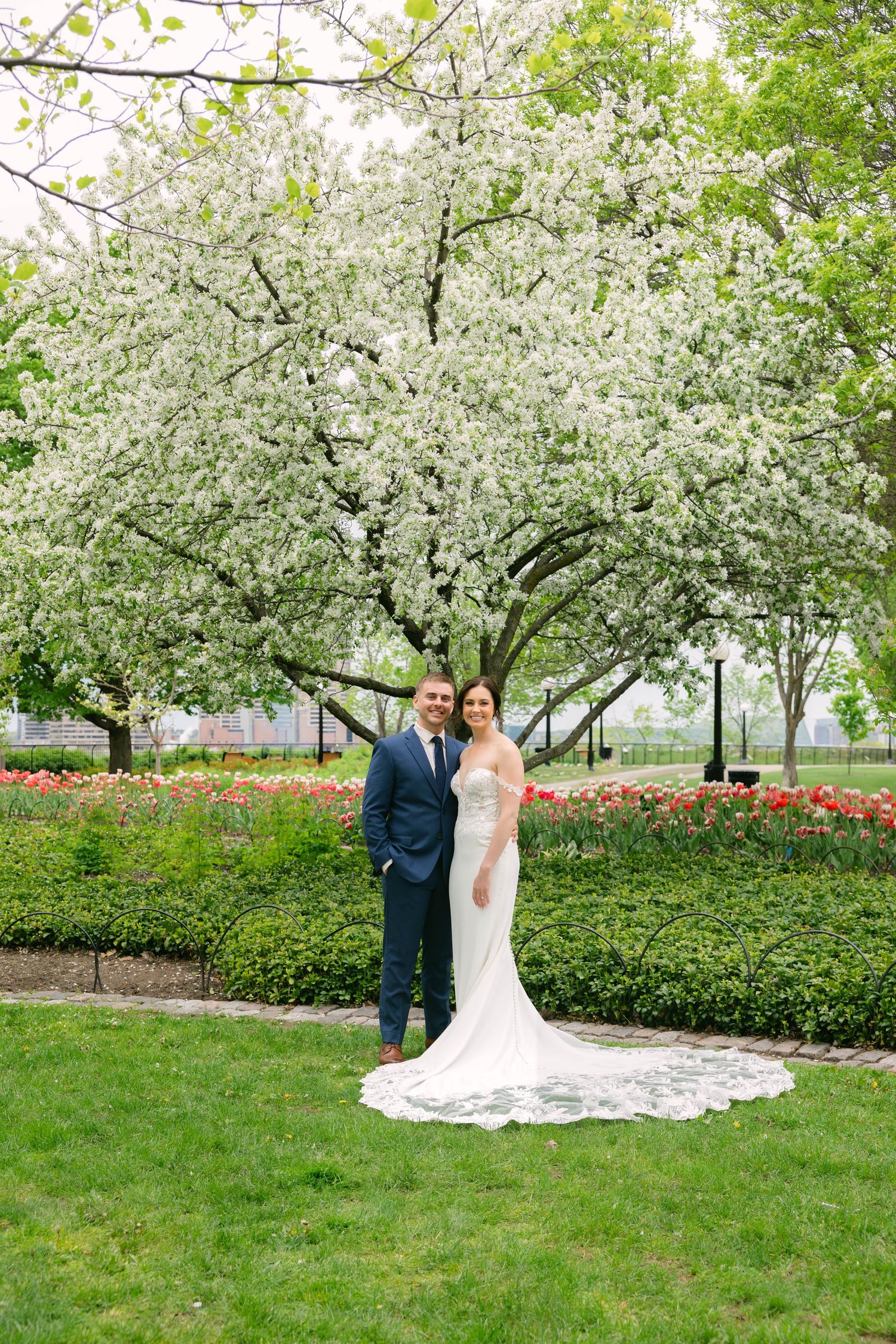 Bride and groom posing in front of a blooming white cherry blossom tree in a lush green garden with tulips.