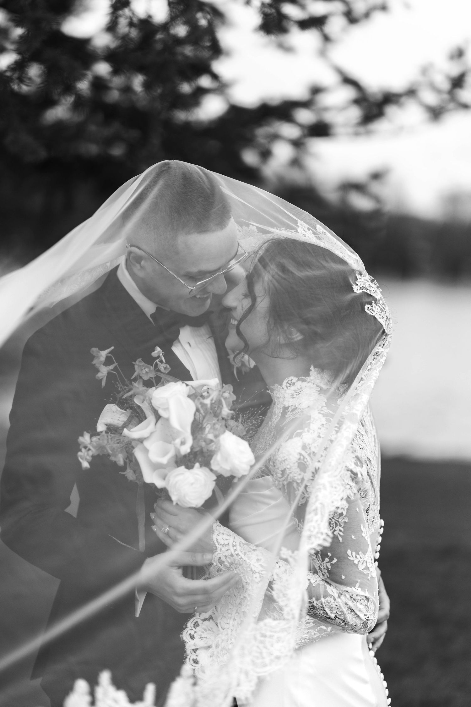 Black and white photo of a bride and groom kissing under a lace veil, bride holding a bouquet outdoors.