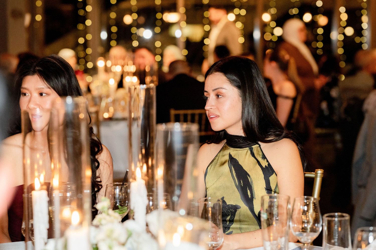 Two elegant women seated at a candlelit gala dinner table with floral centerpieces and warm bokeh lights.