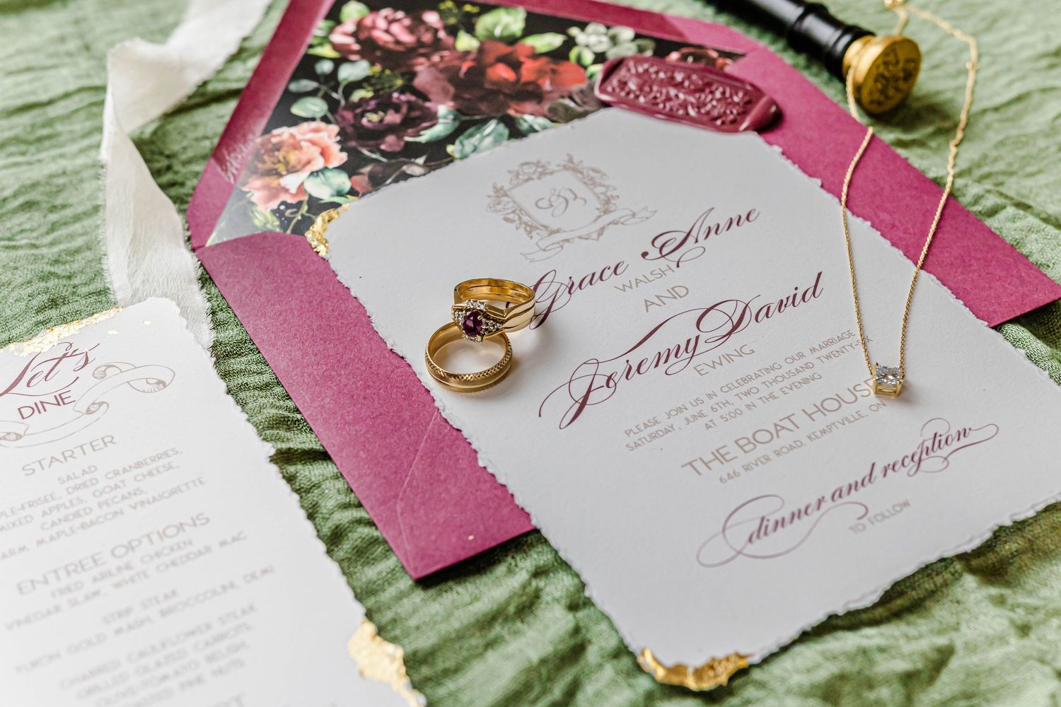 Wedding invitation suite with pink envelope, gold rings, and calligraphy on white paper against green fabric.