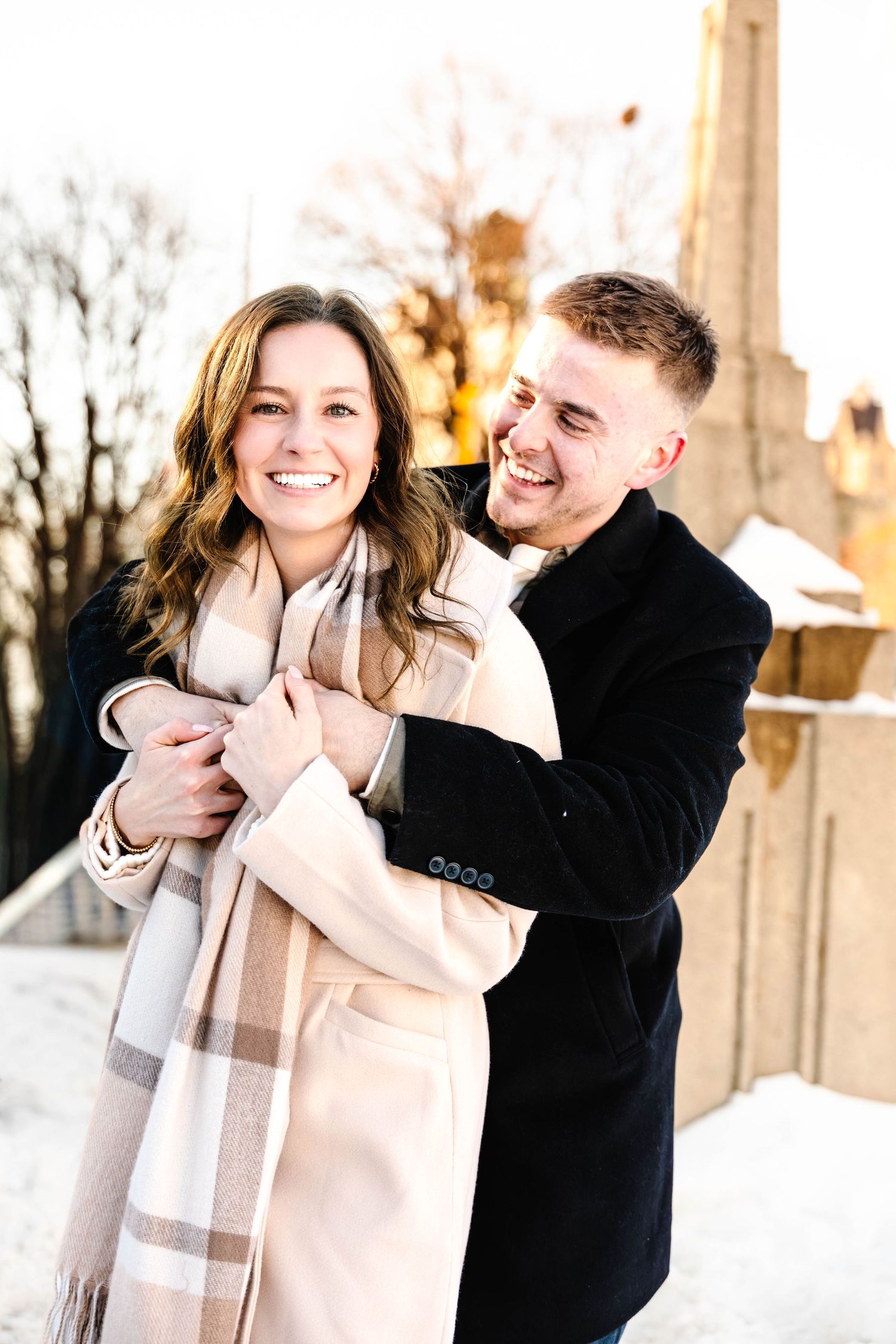 A romantic winter photoshoot captures a couple laughing and embracing in front of snowy trees at sunset.