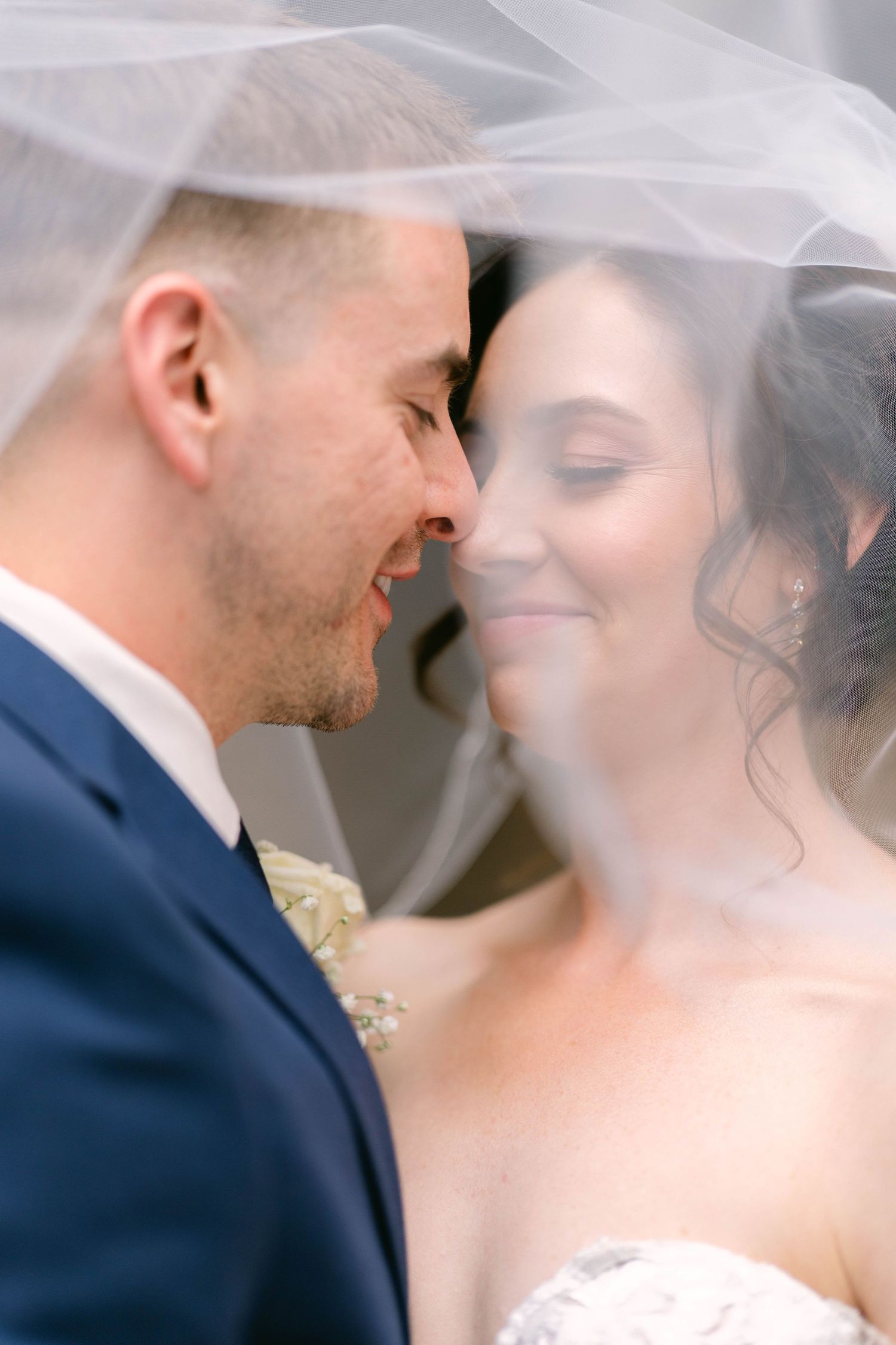 Bride and groom share an intimate moment under a flowing white veil, foreheads touching and smiling softly.