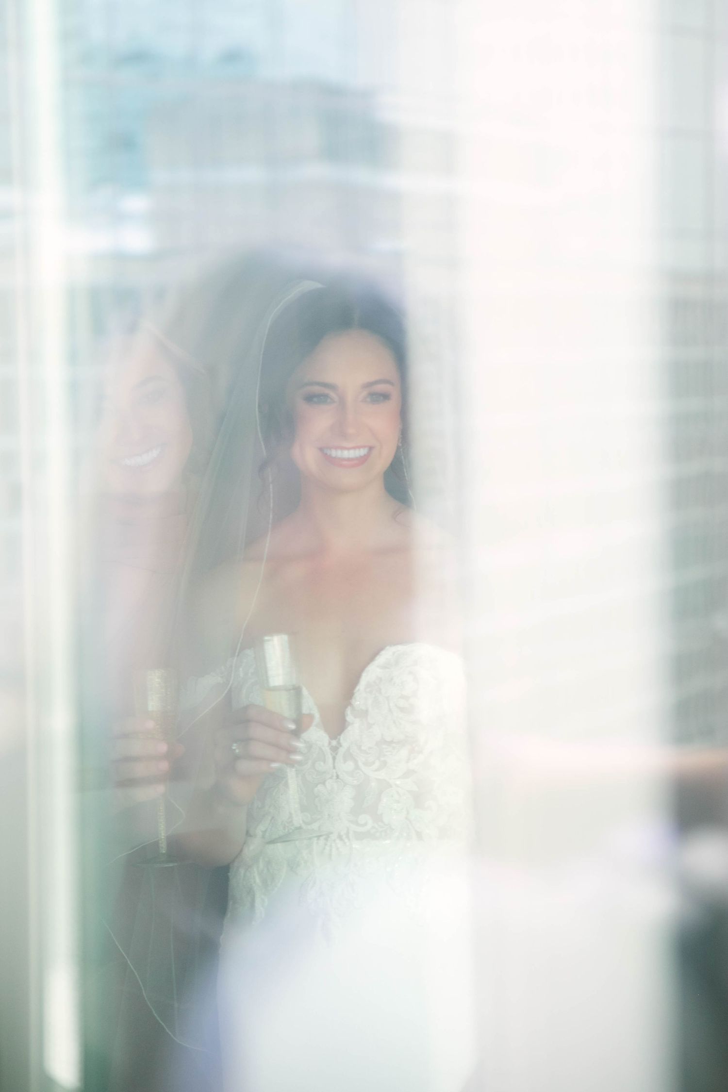 Smiling bride in lace wedding dress and veil holding champagne glass, reflected in window light.