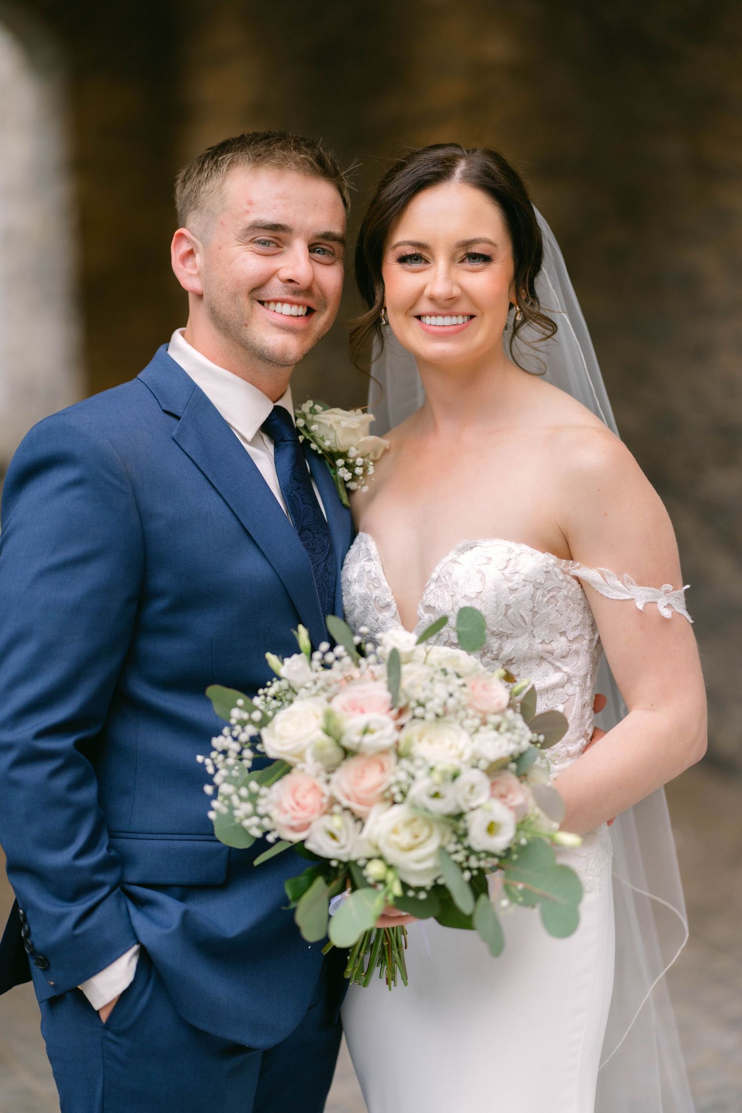 Happy couple on wedding day, bride holding pink and white bouquet in lace off-shoulder gown with veil, groom in navy suit.