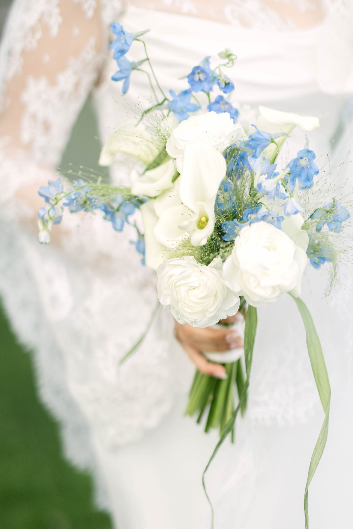 Bridal bouquet of white calla lilies, ranunculus, and blue delphinium flowers held by bride in lace gown.