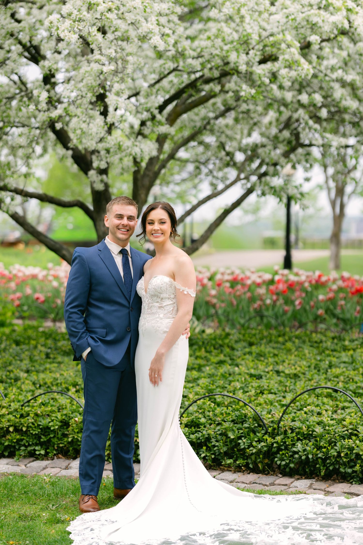 Happy bride and groom pose together in spring garden with white flowering tree and tulips.