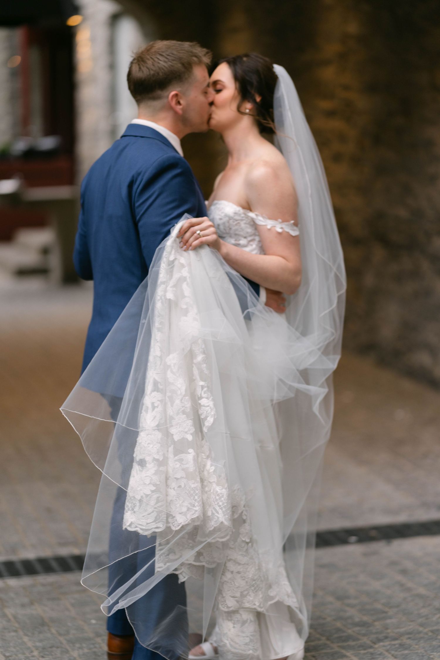 A bride and groom share a romantic kiss, she wears a lace off-shoulder gown with veil, he wears a blue suit.