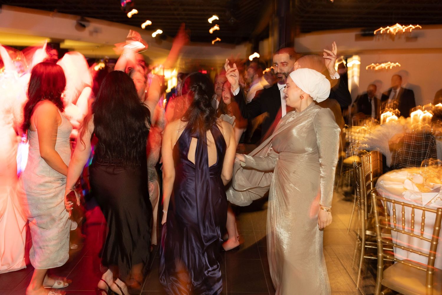 Guests dancing and celebrating at an elegant wedding reception with warm lighting and decorated dining tables.