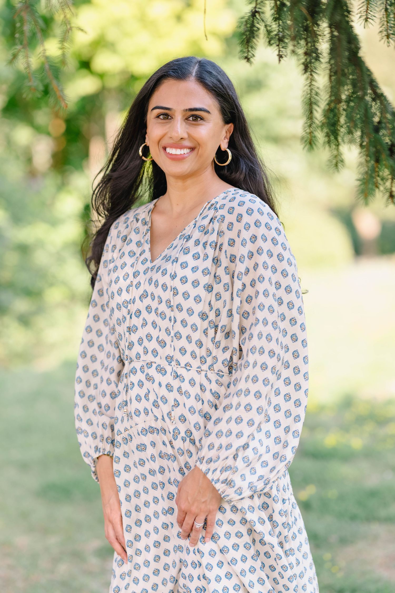 Smiling woman in patterned white dress with gold hoop earrings standing outdoors in a lush green park.