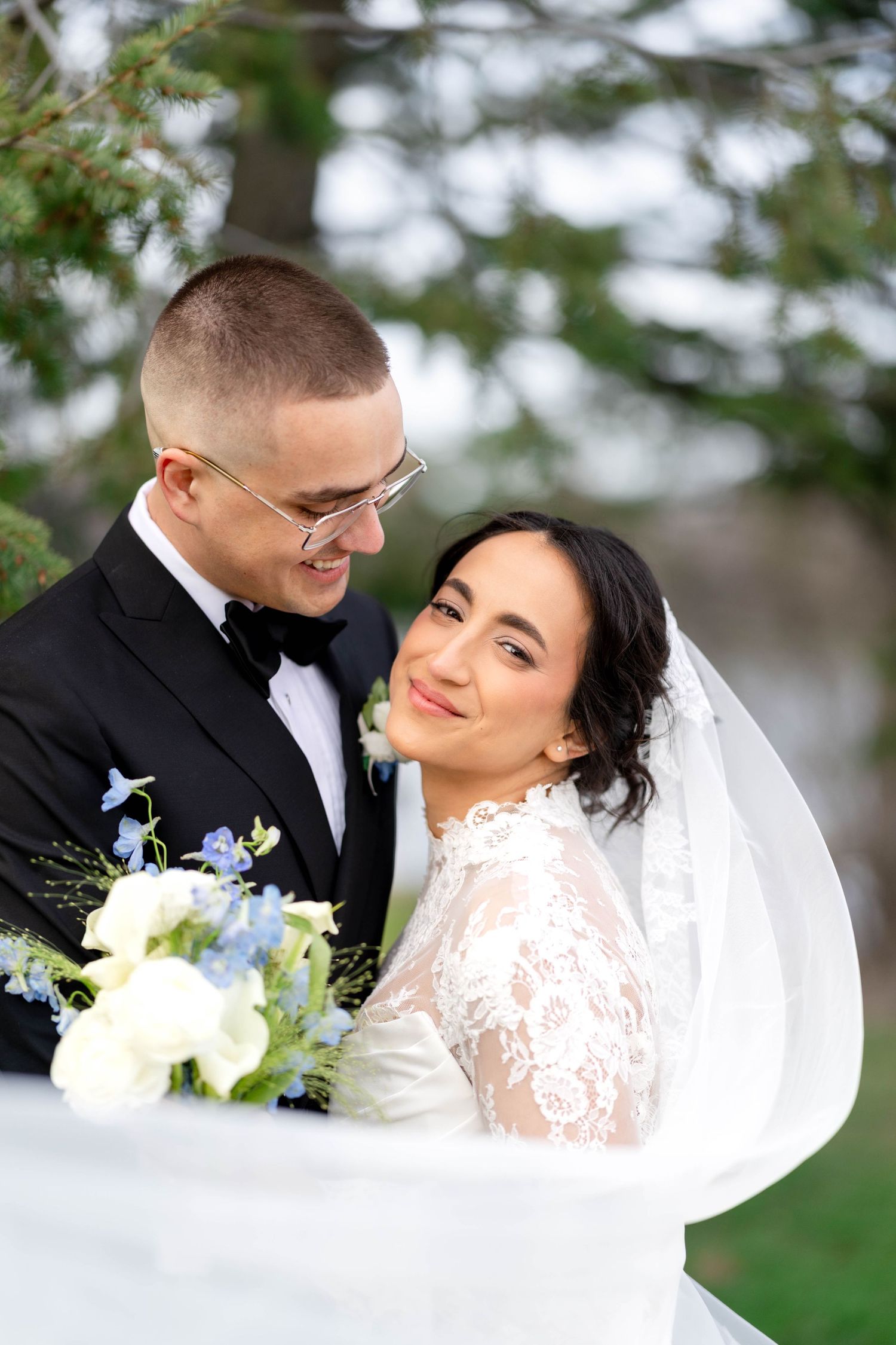 Bride in lace gown and groom in tuxedo embracing under a veil with a blue floral bouquet.