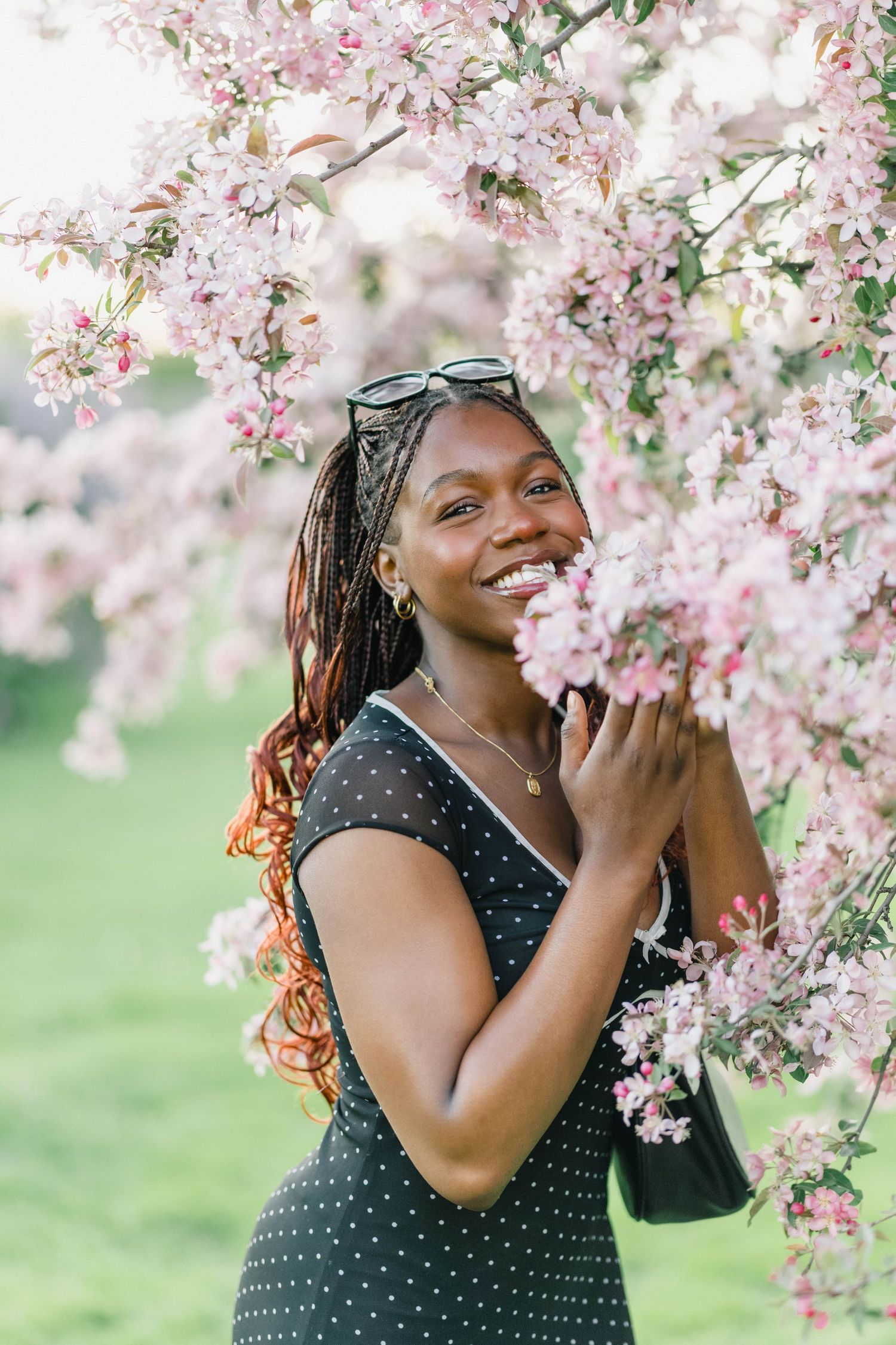 Smiling Black woman in polka dot dress holding pink cherry blossoms in spring garden.