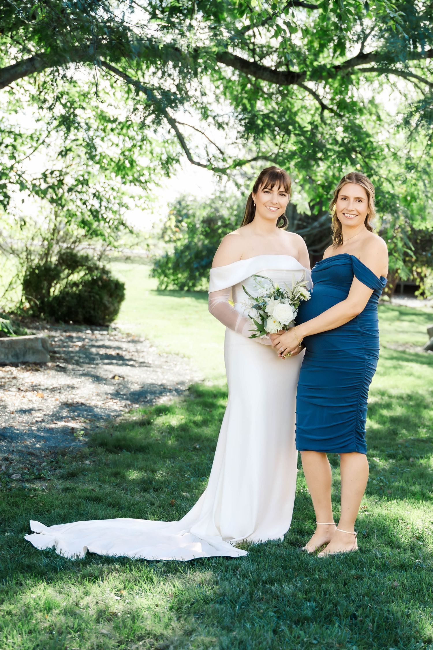 Two women in white and blue dresses share a playful moment together on green grass in a sunny outdoor setting.