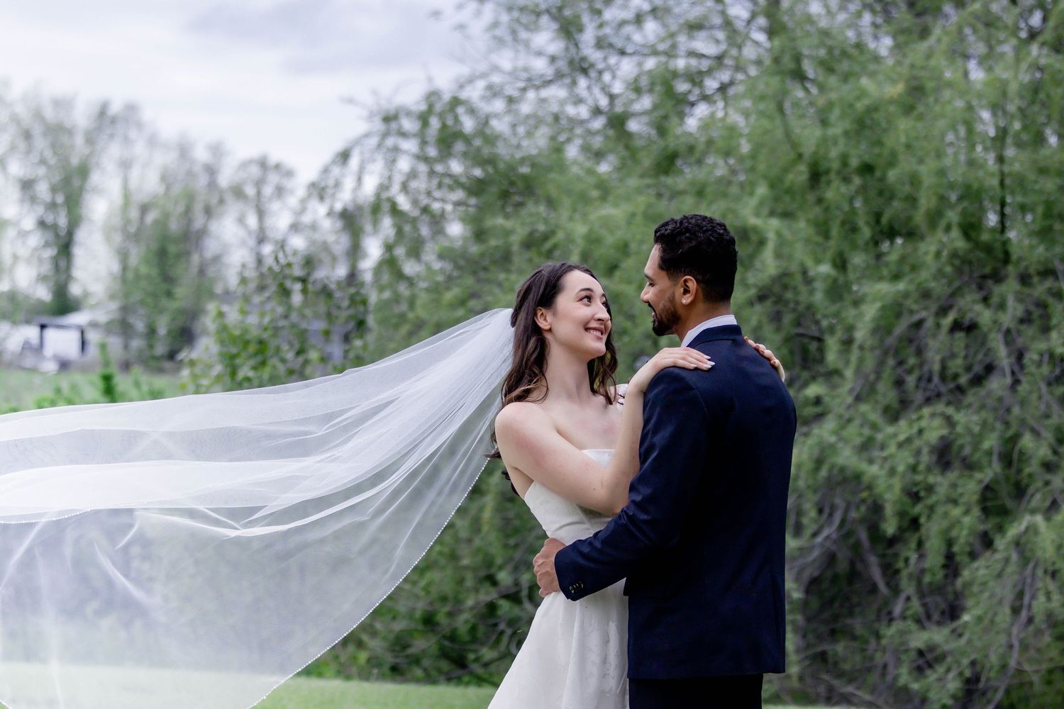 A couple in wedding attire dances together as a flowing veil creates a romantic silhouette against a natural backdrop.