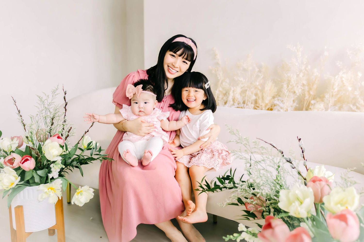 Family in matching pink dresses poses on couch surrounded by elegant floral arrangements and soft lighting.