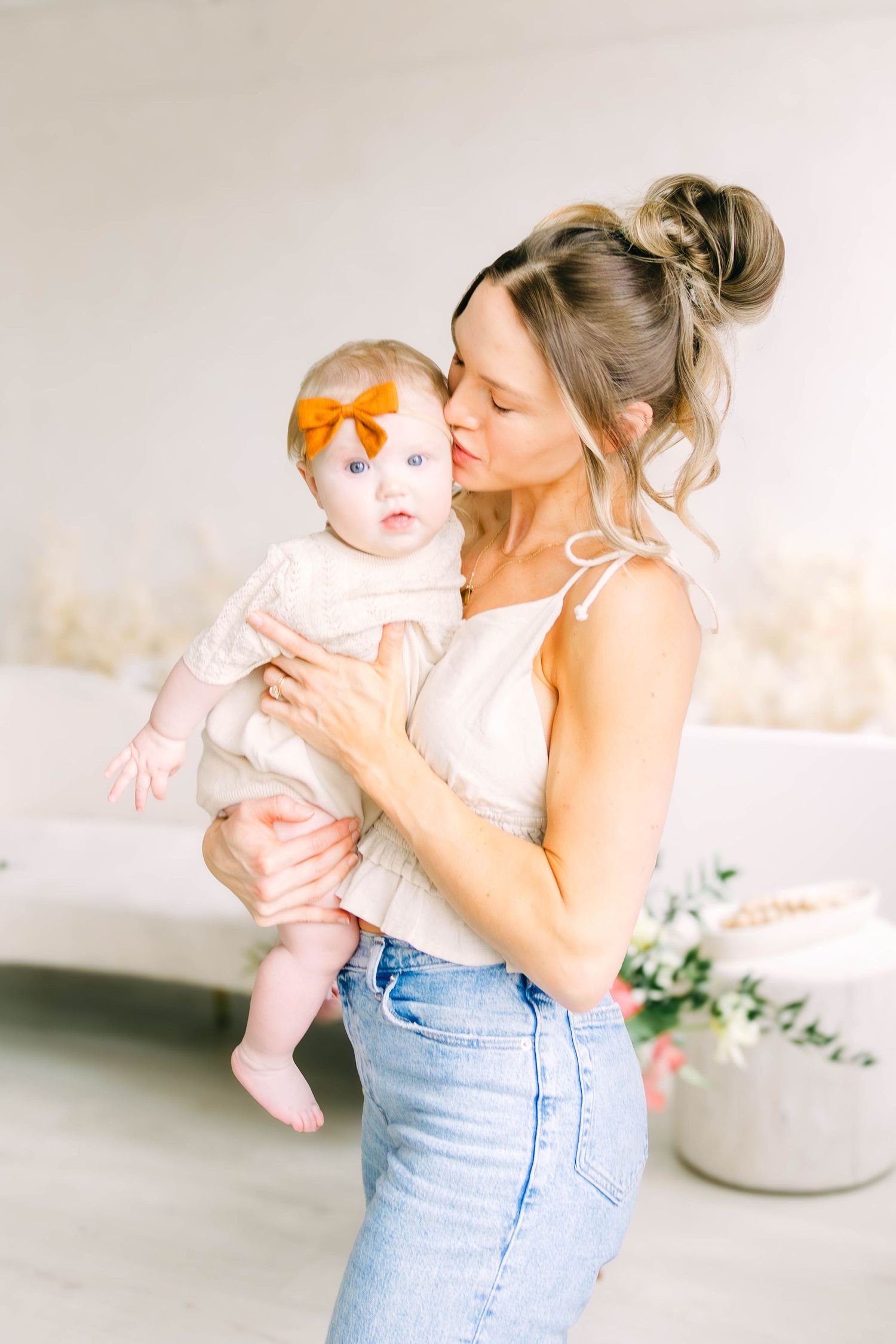 Mother in blue jeans holds and kisses baby in white outfit against light background in sweet candid photo series.