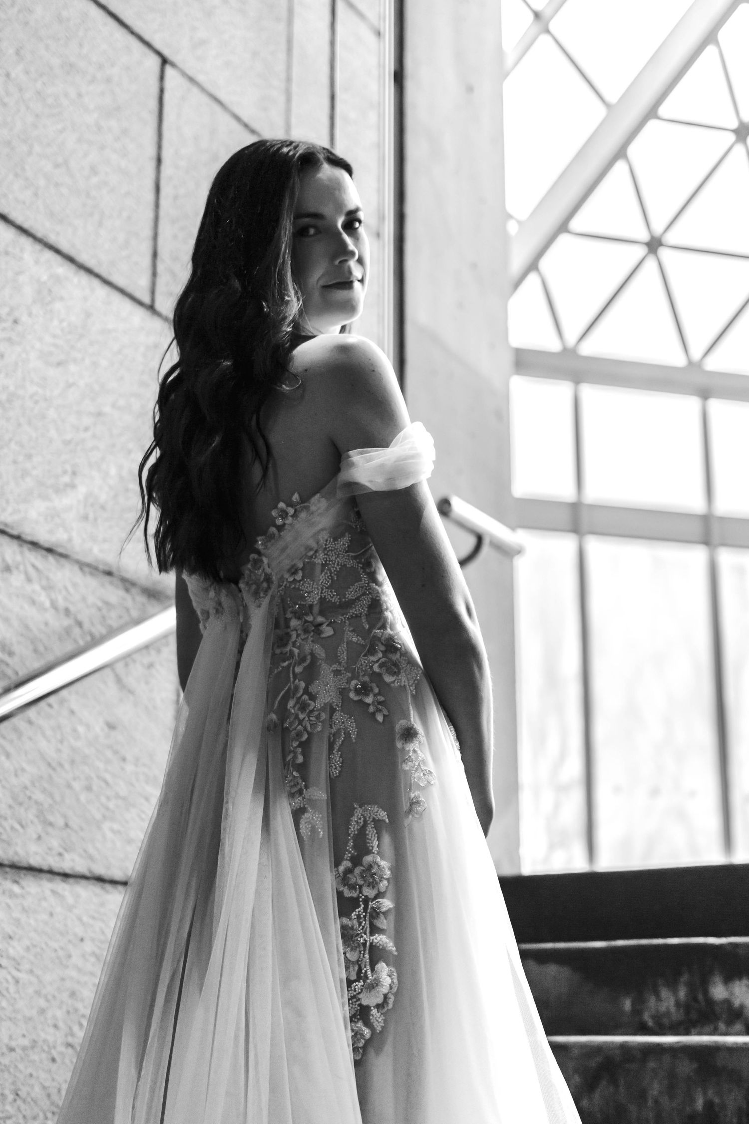 A bride in an elegant white lace wedding dress poses in a sunlit room with large windows.