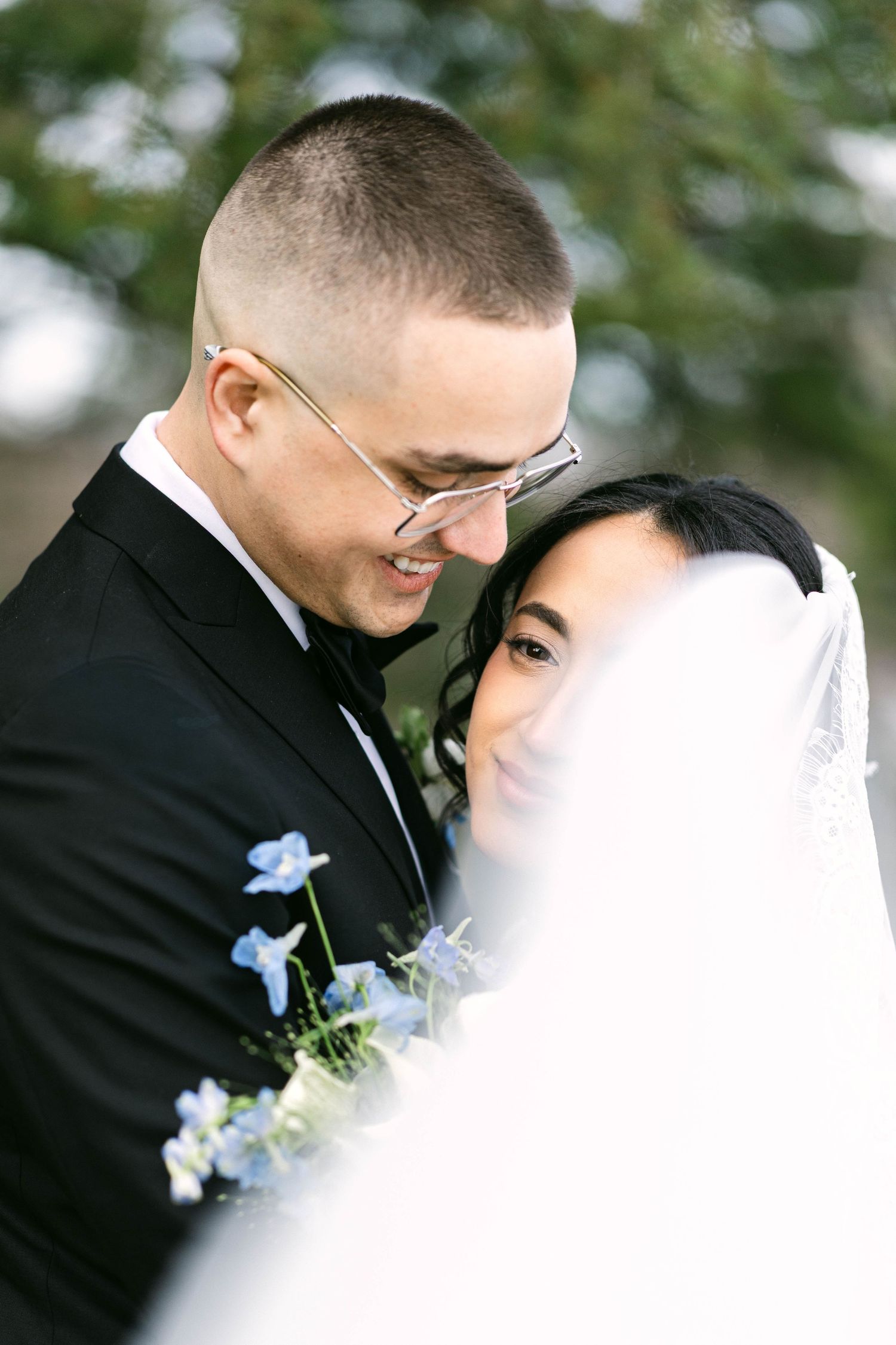 Bride and groom share tender moment, groom in black tuxedo with glasses, bride in white veil with blue floral accents.