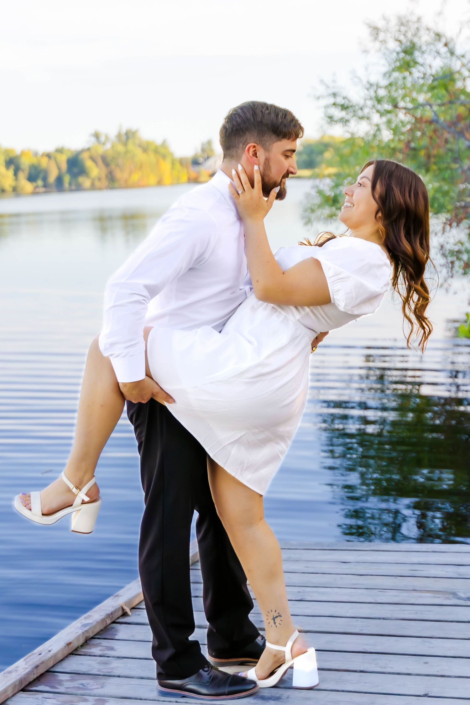 Couple in contrasting outfits dance playfully on a lakeside dock during golden hour.