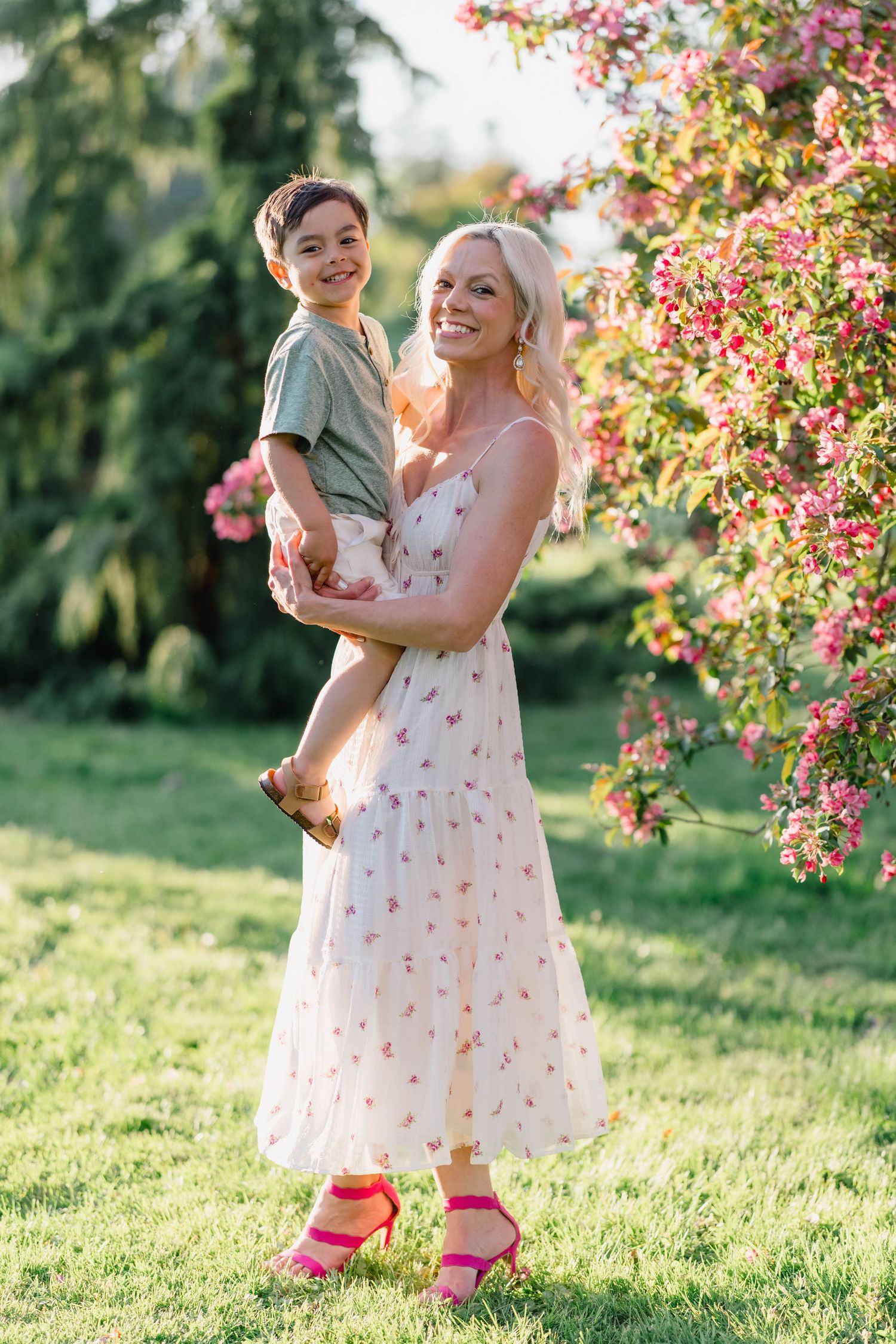 Smiling blonde woman in floral dress holds young boy near blooming pink flowering tree.