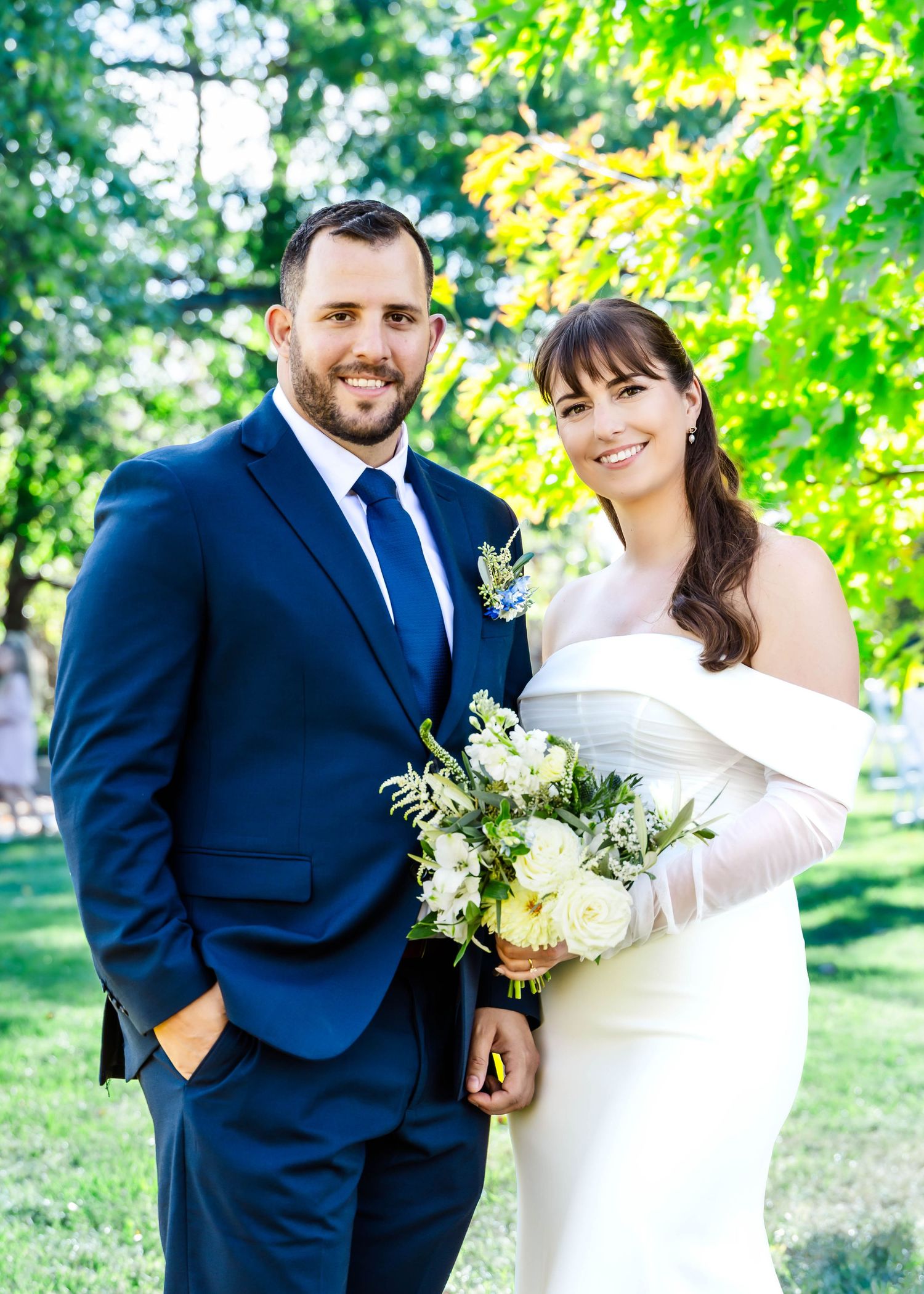A couple in wedding attire smiles while holding a white floral bouquet in a garden setting.