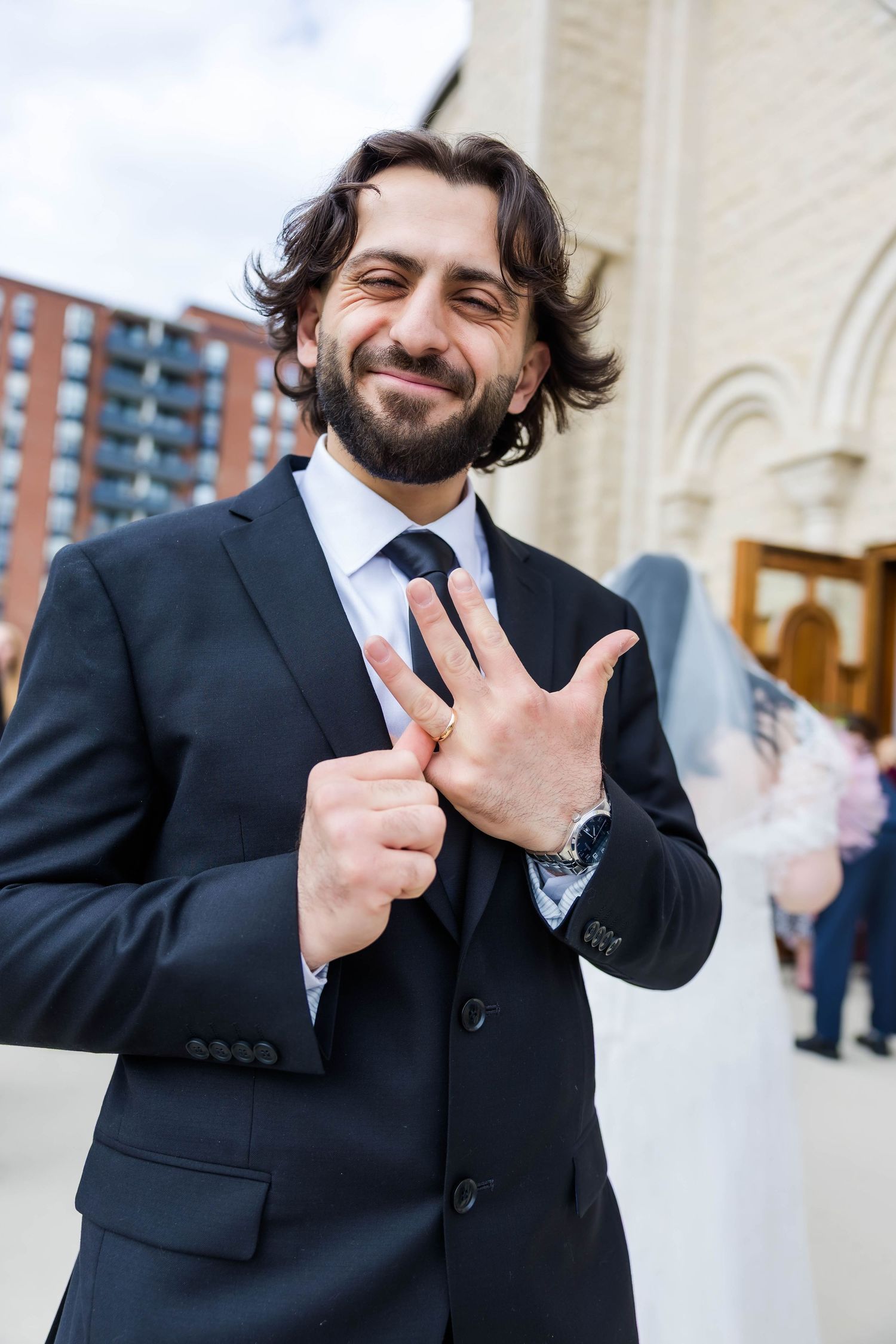 A well-dressed person in a dark suit and tie gestures happily outside a building with architectural details.