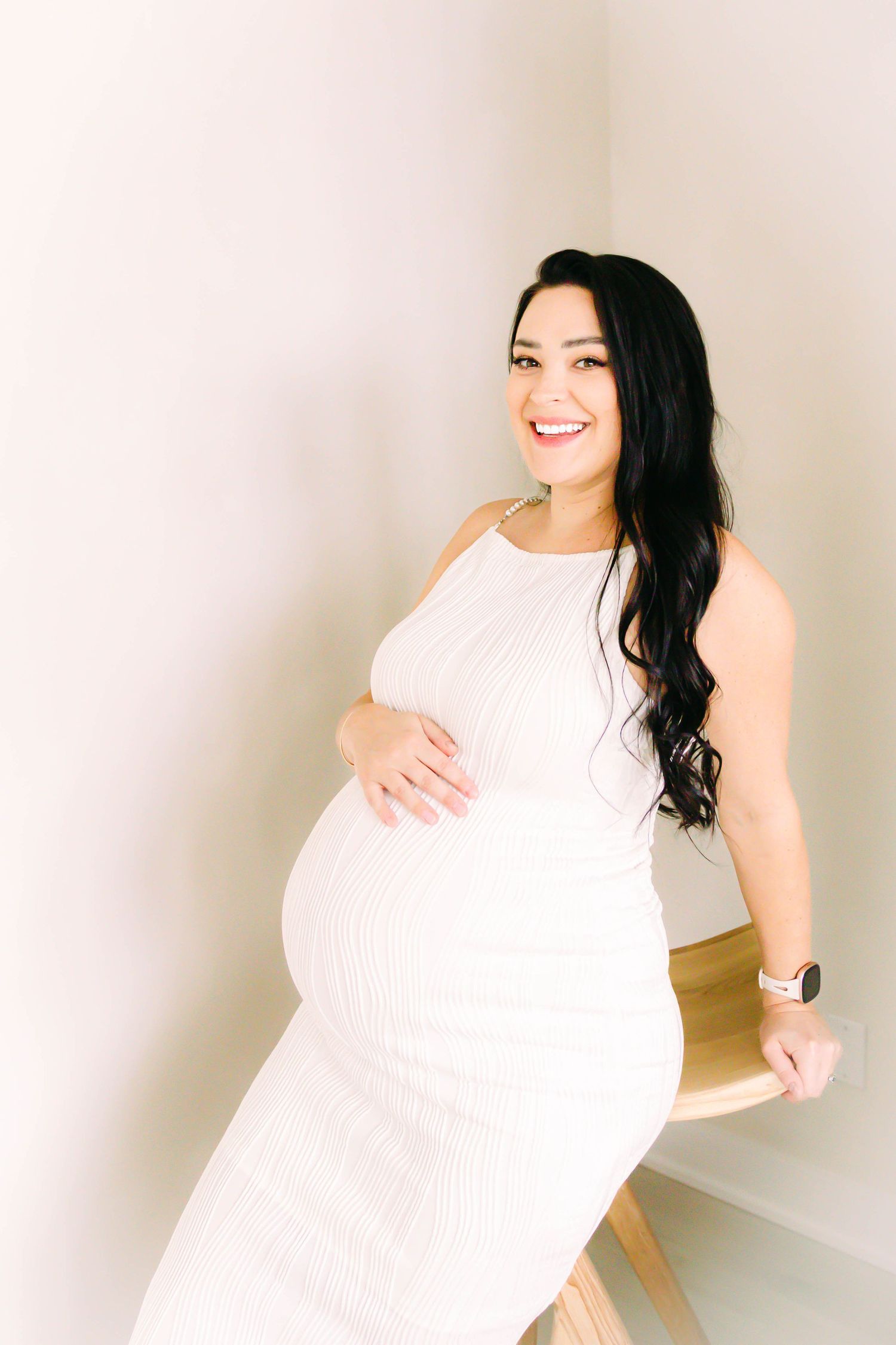 A person in a white ribbed dress poses during a maternity photo shoot against a light colored wall.
