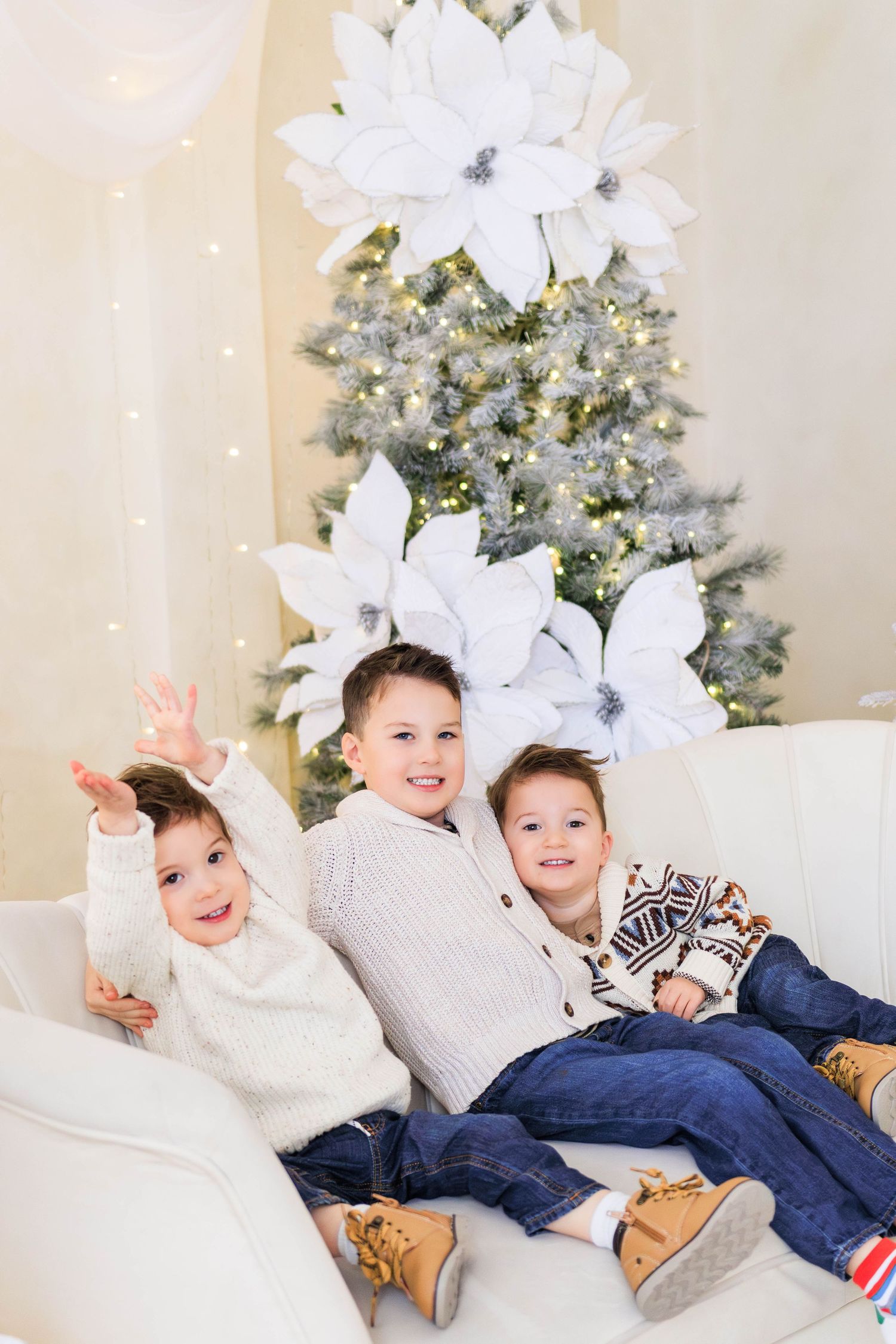Three children pose playfully on a white couch with decorated Christmas trees in the background.