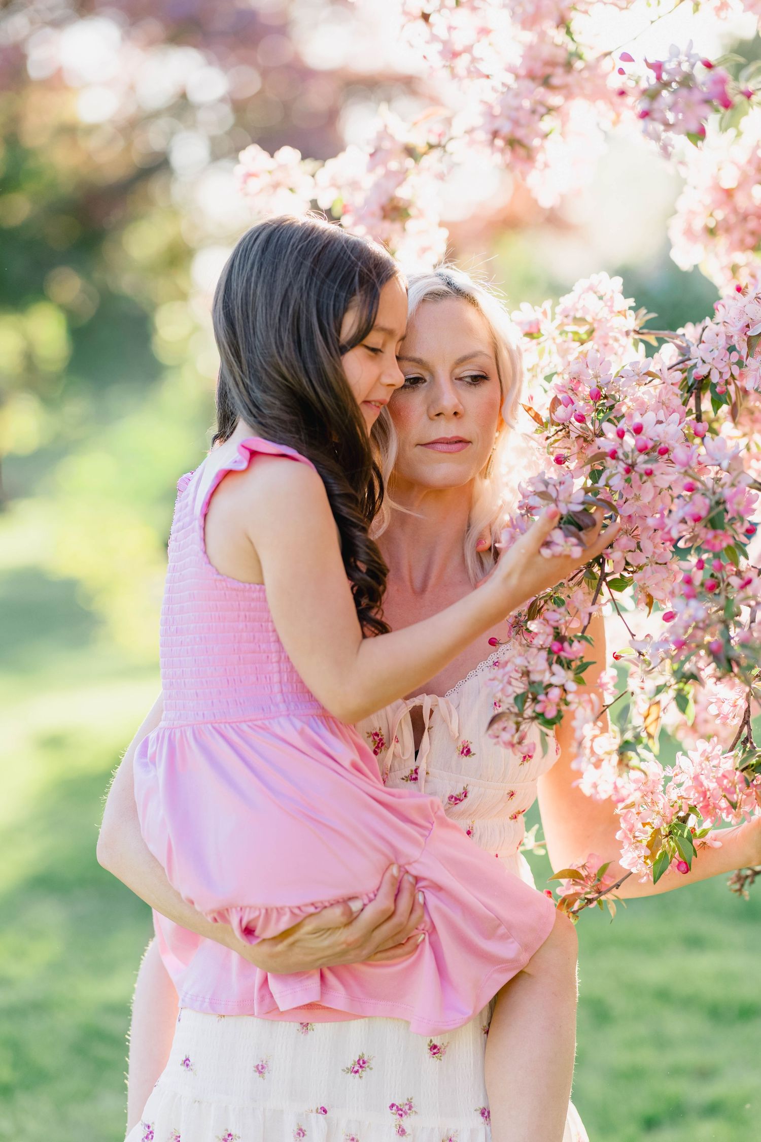 Mother holds young daughter near blooming pink cherry blossom tree in a sunlit garden.