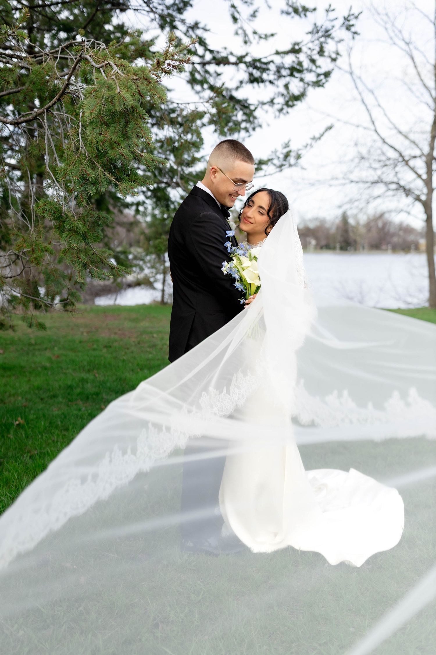 Bride and groom embrace outdoors with flowing lace veil billowing in wind, standing near lake in wedding attire.