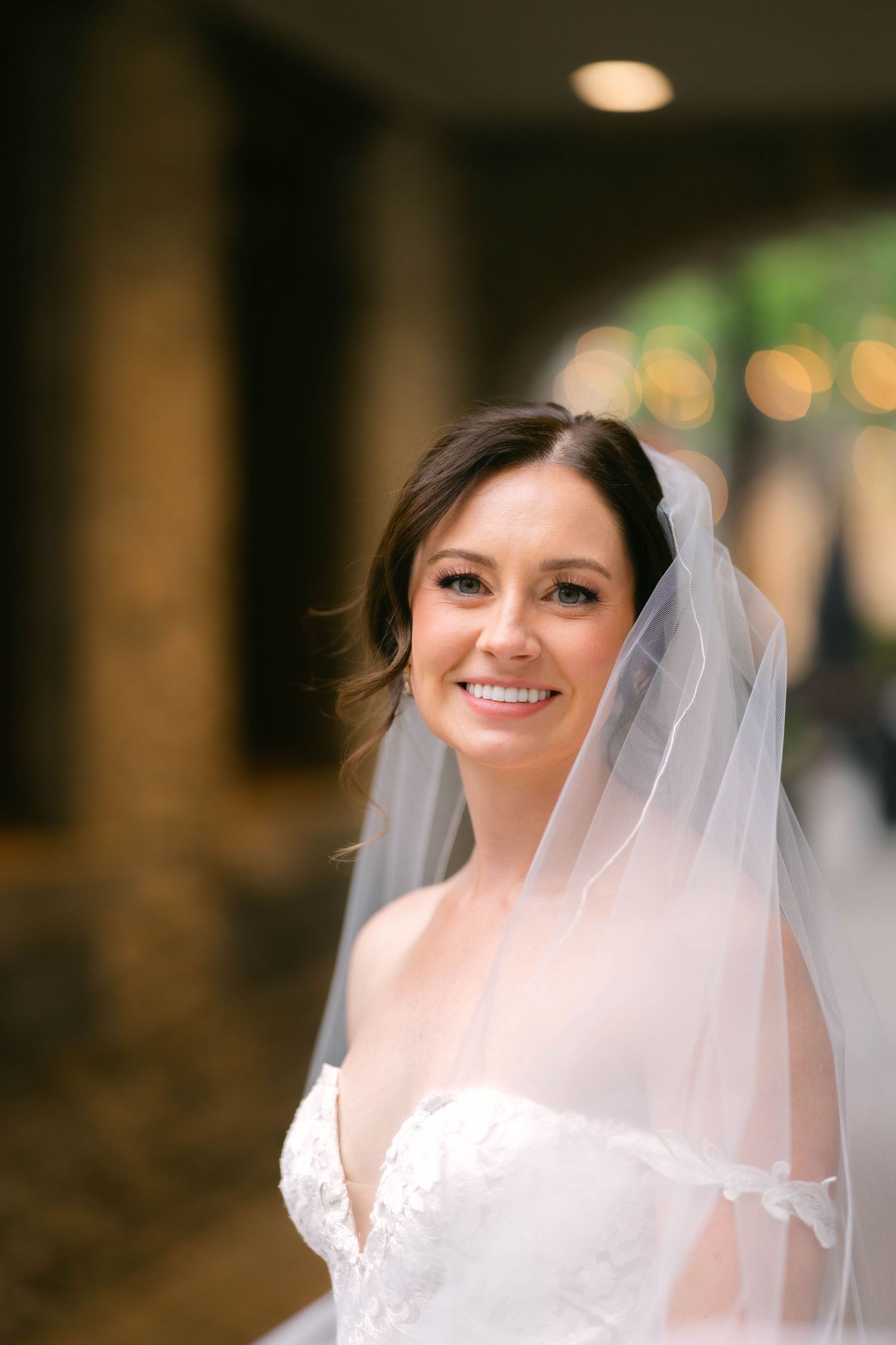 Smiling bride in lace gown poses elegantly with veil draped over her shoulders.