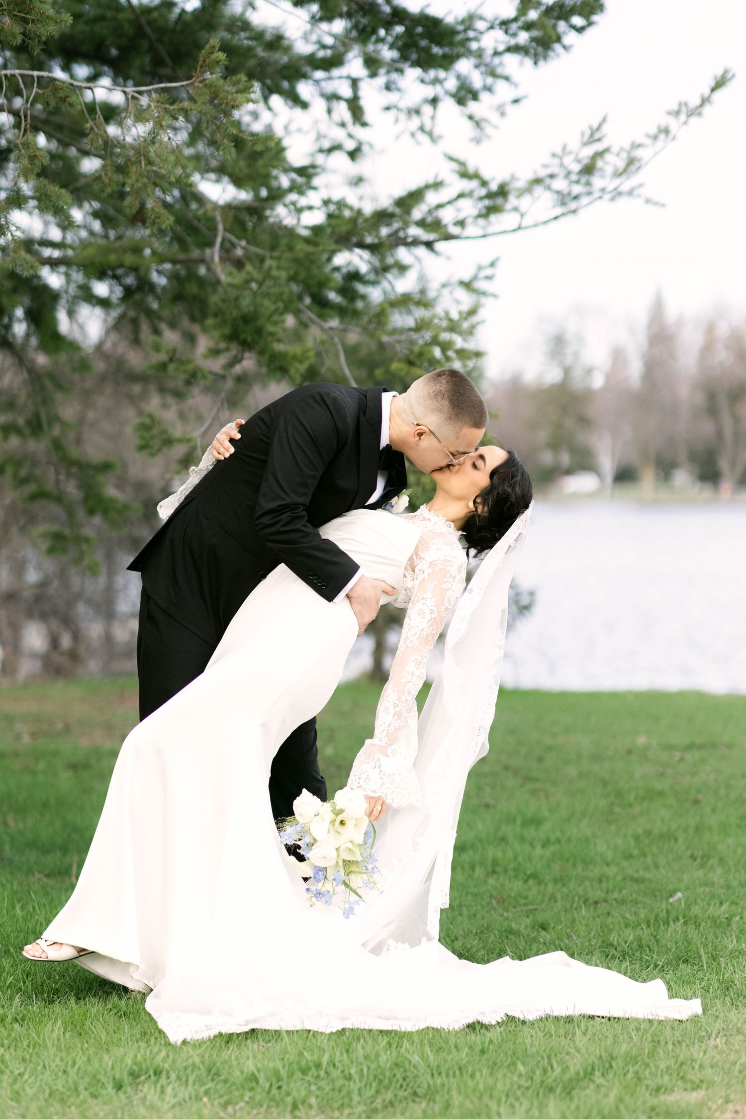 Groom in black tuxedo dips bride in lace wedding gown for a romantic kiss on a green lawn near a lake.