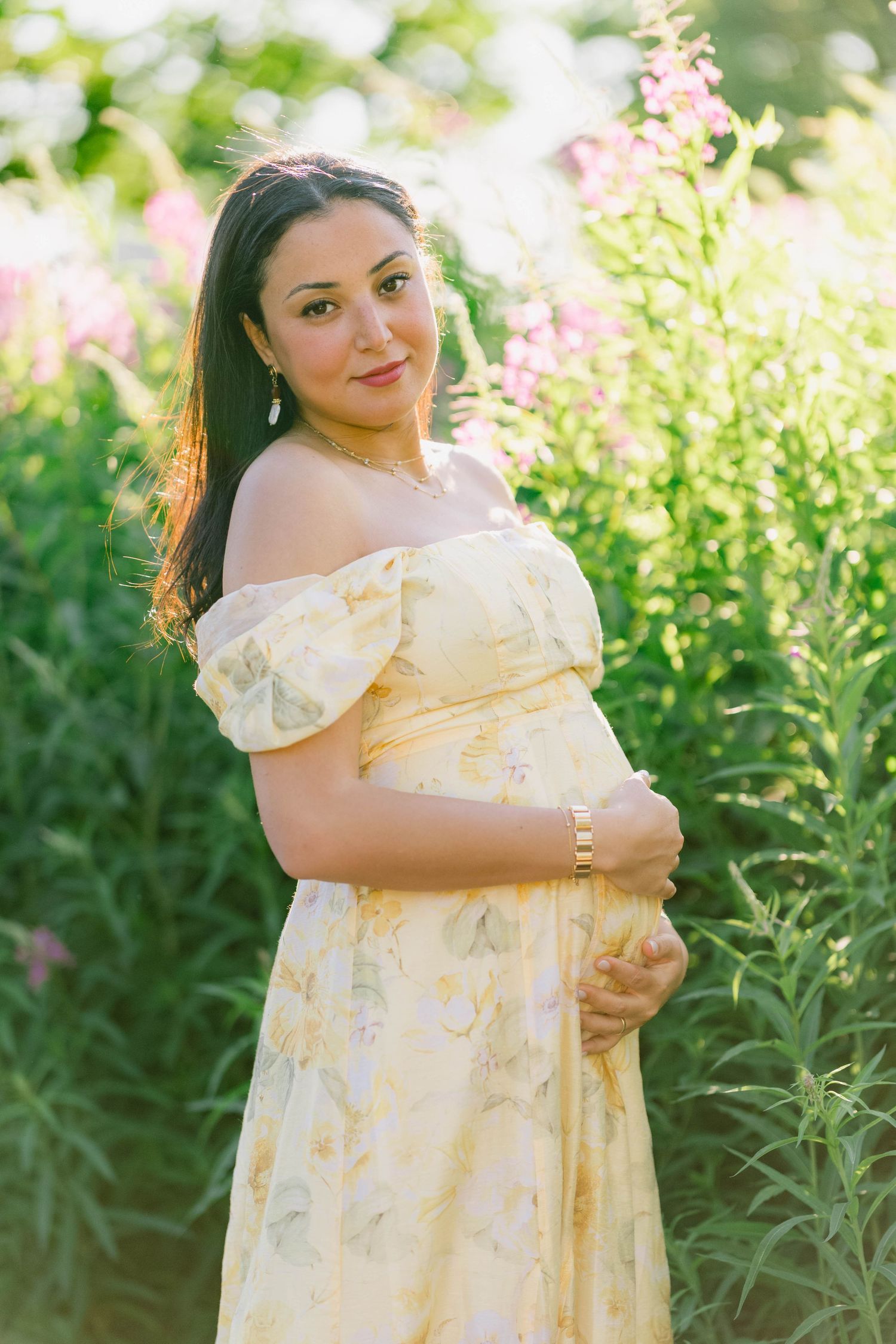 Pregnant woman in yellow floral off-shoulder dress smiling outdoors among pink wildflowers in golden sunlight.