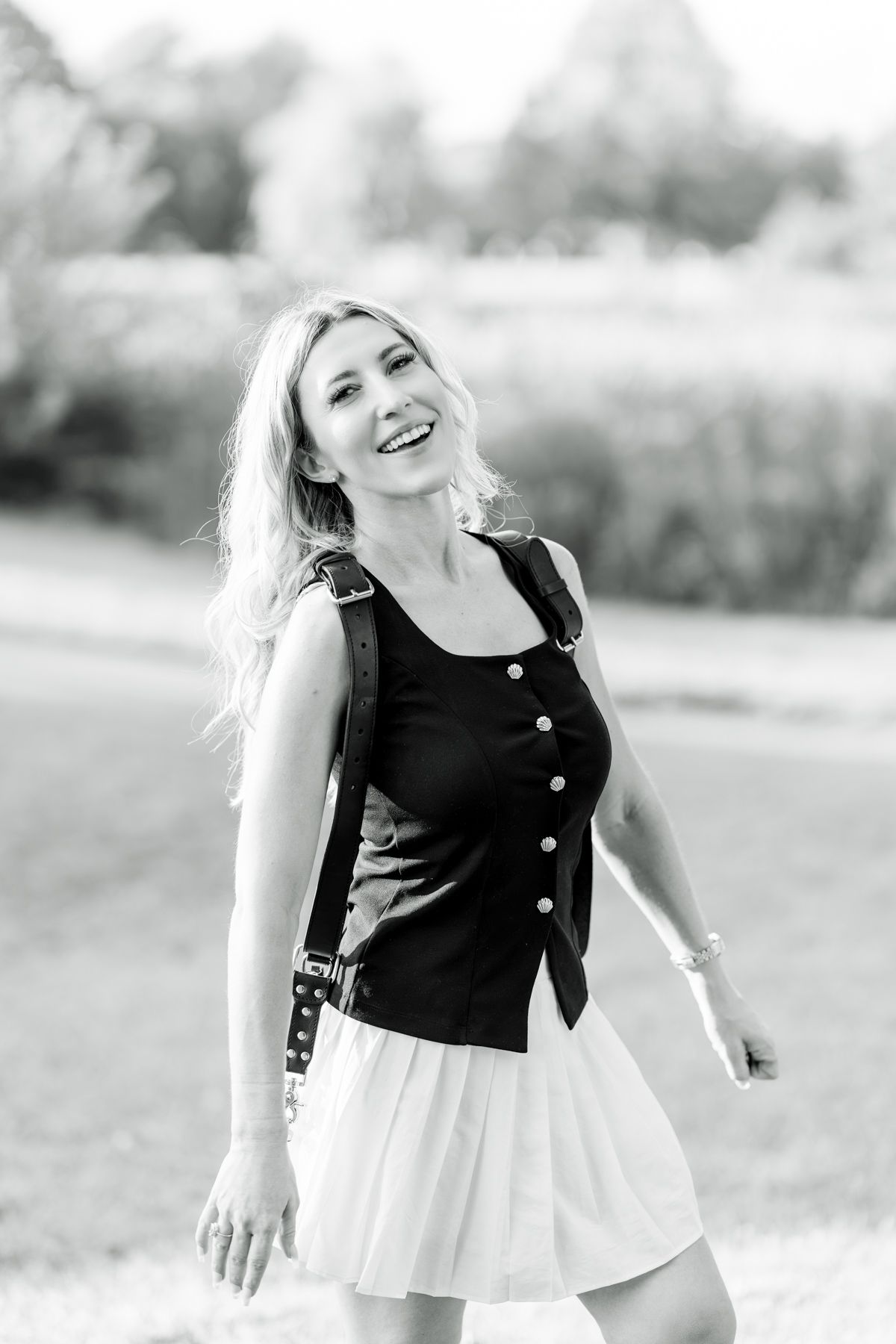 Black and white photo of a smiling blonde woman in a black top and white skirt, posing outdoors in a park.