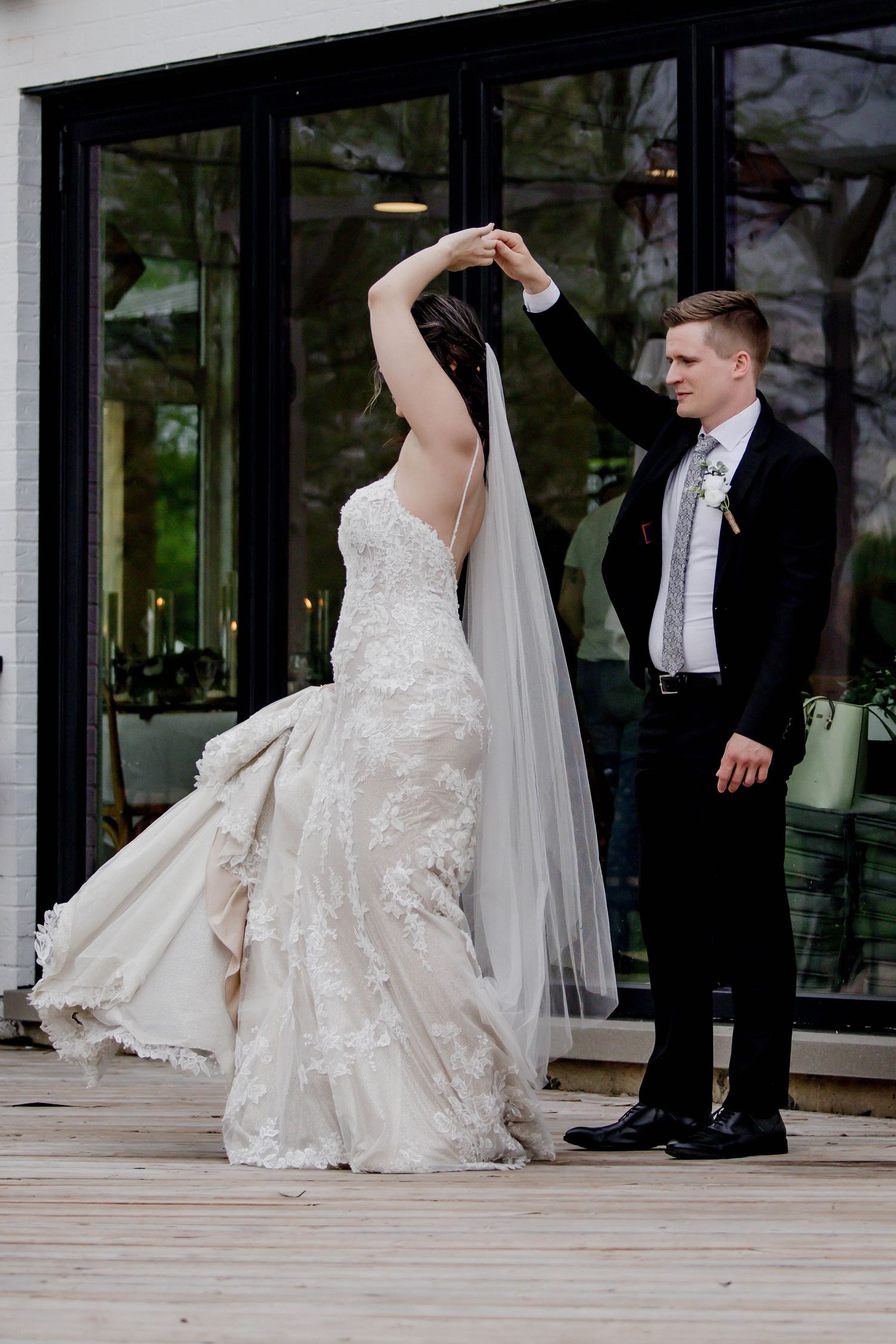 A bride in a white lace gown and groom in a black tuxedo dance together on an outdoor patio during their wedding.
