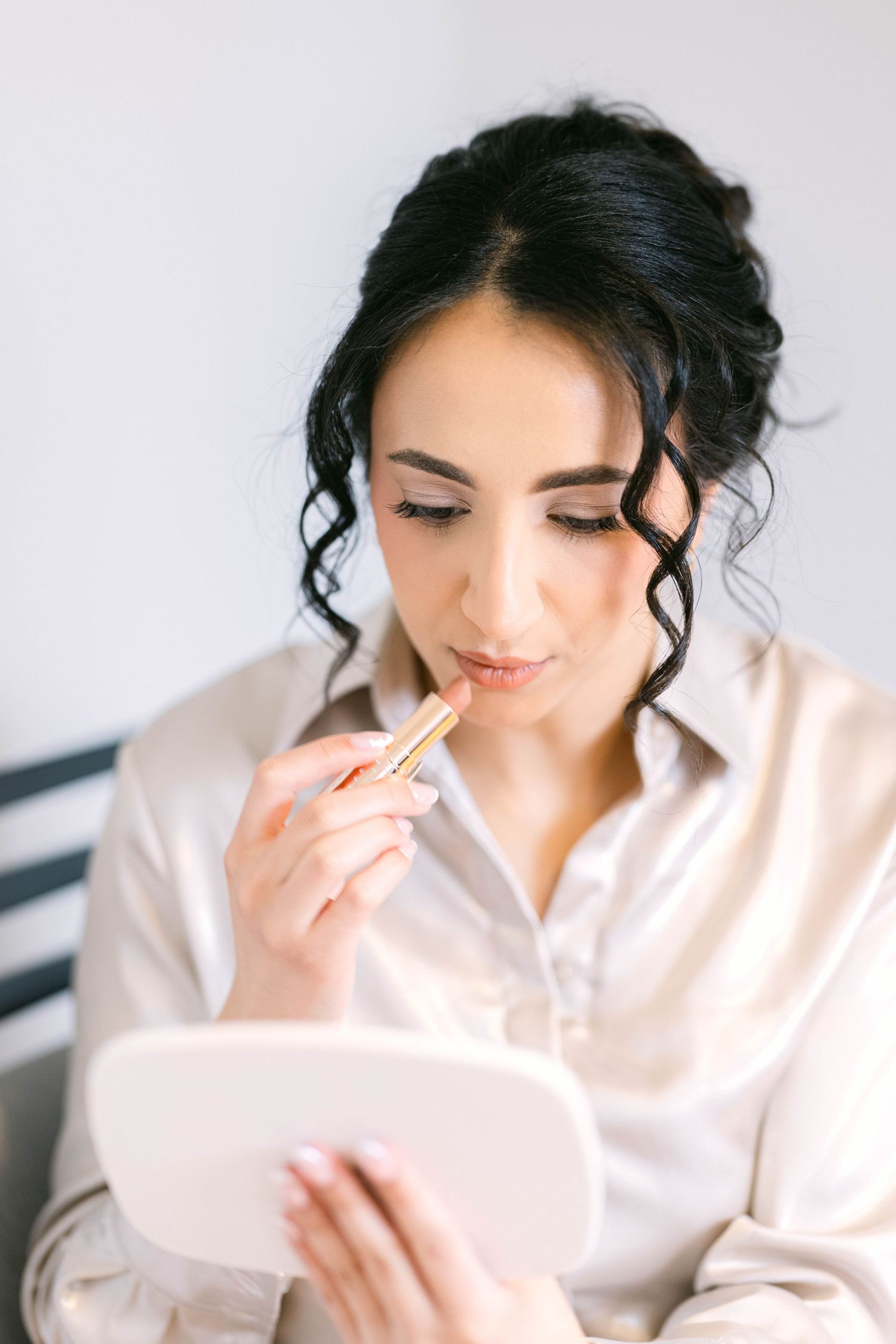 Dark-haired bride applies nude lipstick using a compact mirror while getting ready in a silk blouse.